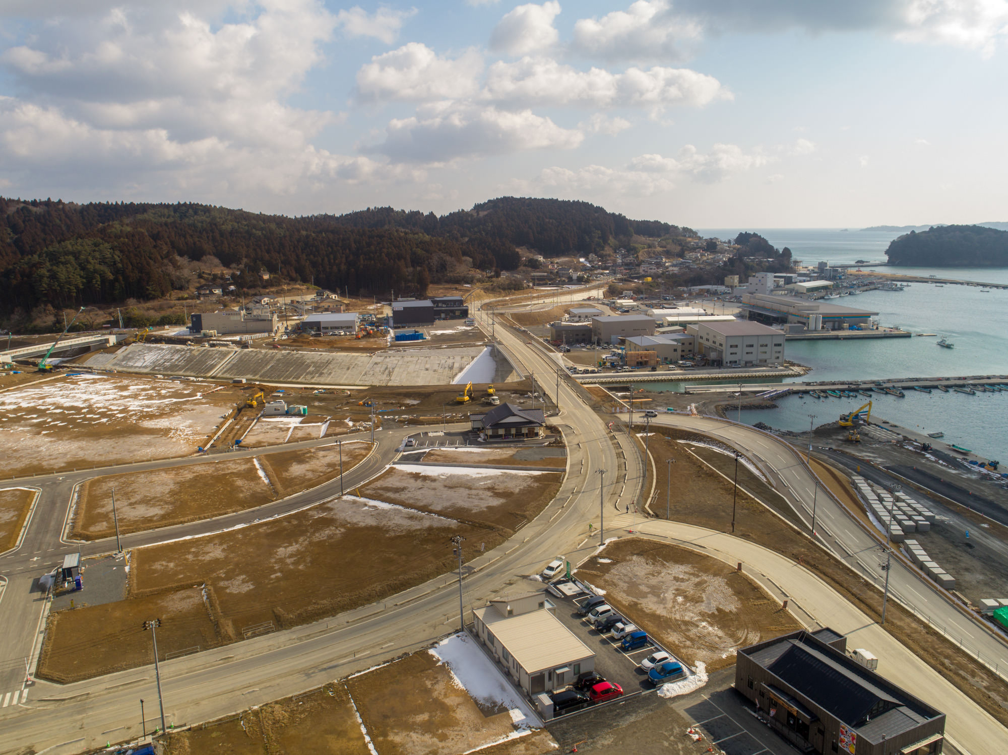 海沿いの地域を上空から撮影した写真で、複数の曲線や直線の道路が交差しながら海岸へと続き、桟橋や水辺が広がり、周囲には建物や工事現場が点在し、地面には一部雪が残り、背景には森林に覆われた丘が広がる風景の写真