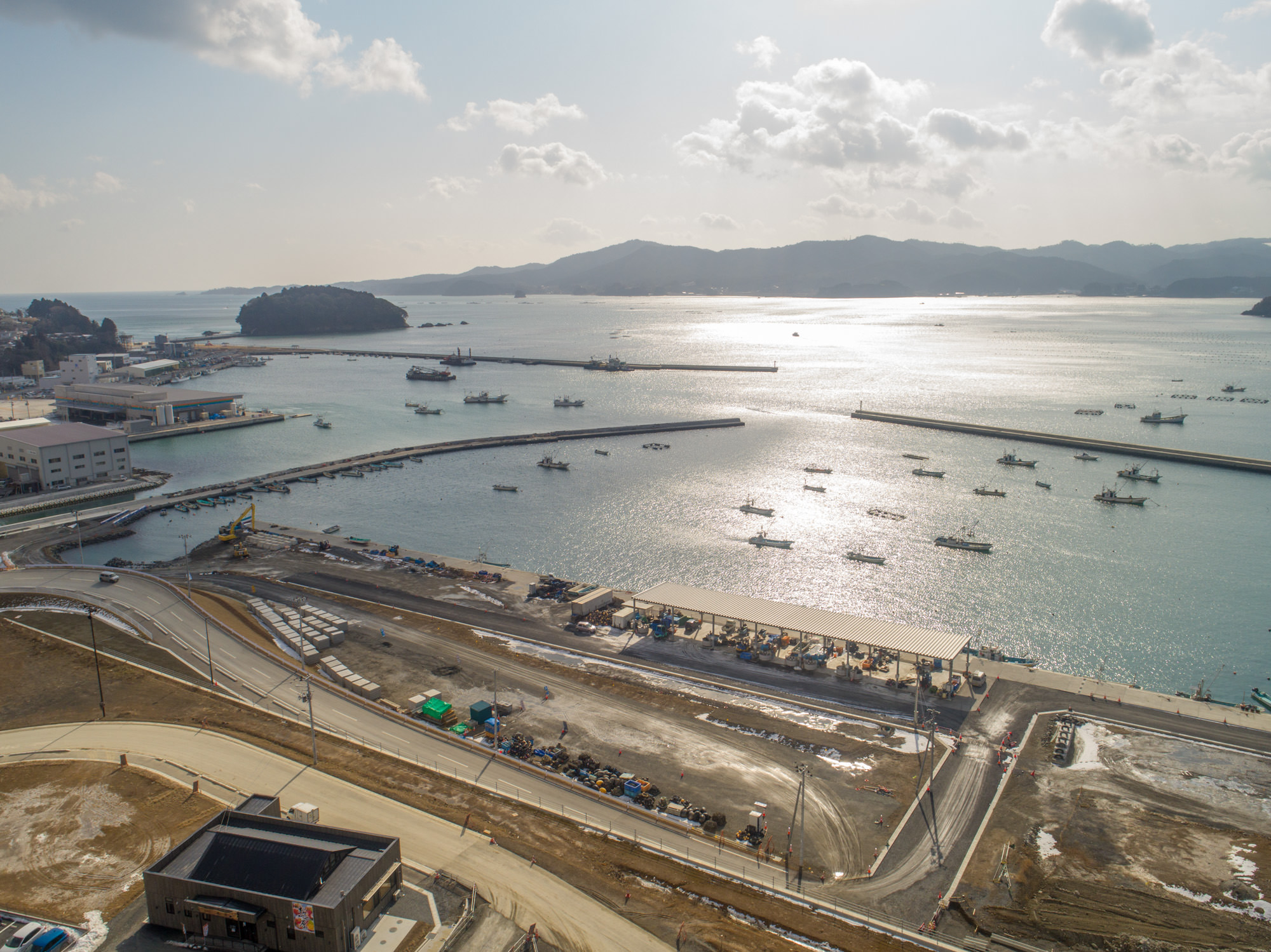 港のある海沿いの地域を上空から撮影した写真で、海には複数の船が浮かび、防波堤によって守られており、海岸沿いには建物や道路、工事中の施設が並び、背景には丘や山が広がり、空は雲が少し浮かび水面に太陽の光が反射する風景の写真