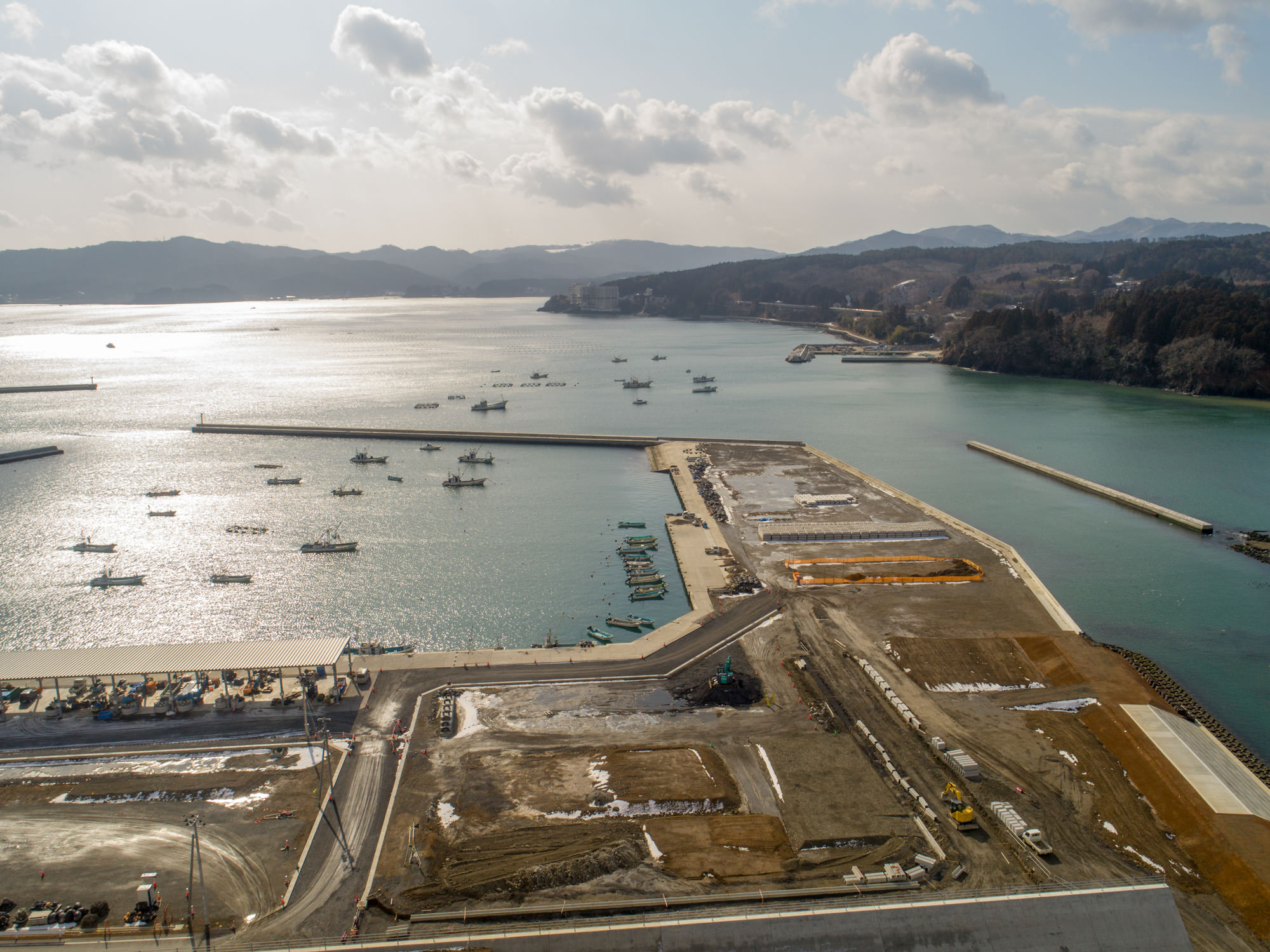 港のある海沿いの地域を上空から撮影した写真で、海には複数の船が停泊し、防波堤が港を囲み、隣接する陸地では工事が進められており、建設機材や資材が見られ、背景には丘や山が広がり、空は雲が少し浮かぶ風景の写真