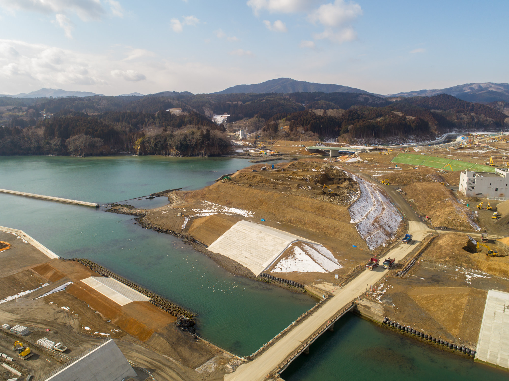 川から海へと続く水域を中心に、周囲の陸地に複数の構造物や建設現場が広がる沿岸地域を上空から撮影したもので、道路や橋、堤防などが確認でき、背景には丘や山々が広がり、地面には雪が残る寒冷地の様子が見られる沿岸の建設風景と自然地形の写真
