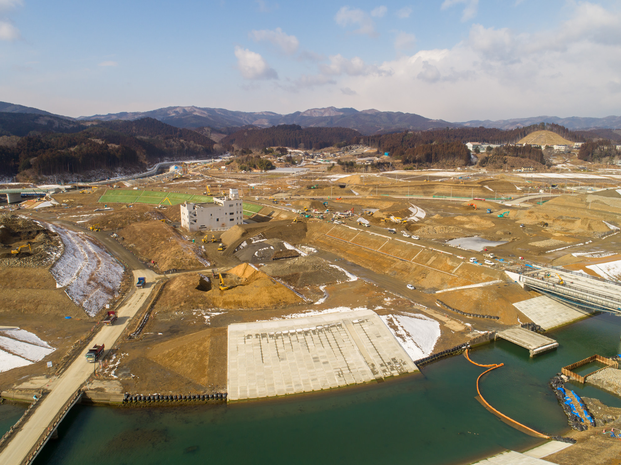 川沿いの近くに広がる大規模な建設現場を写したもので、複数の構造物や重機が点在し、背景には丘や山々が広がり、地面には雪がまだらに残っている様子が見られる建設現場と周辺風景の写真