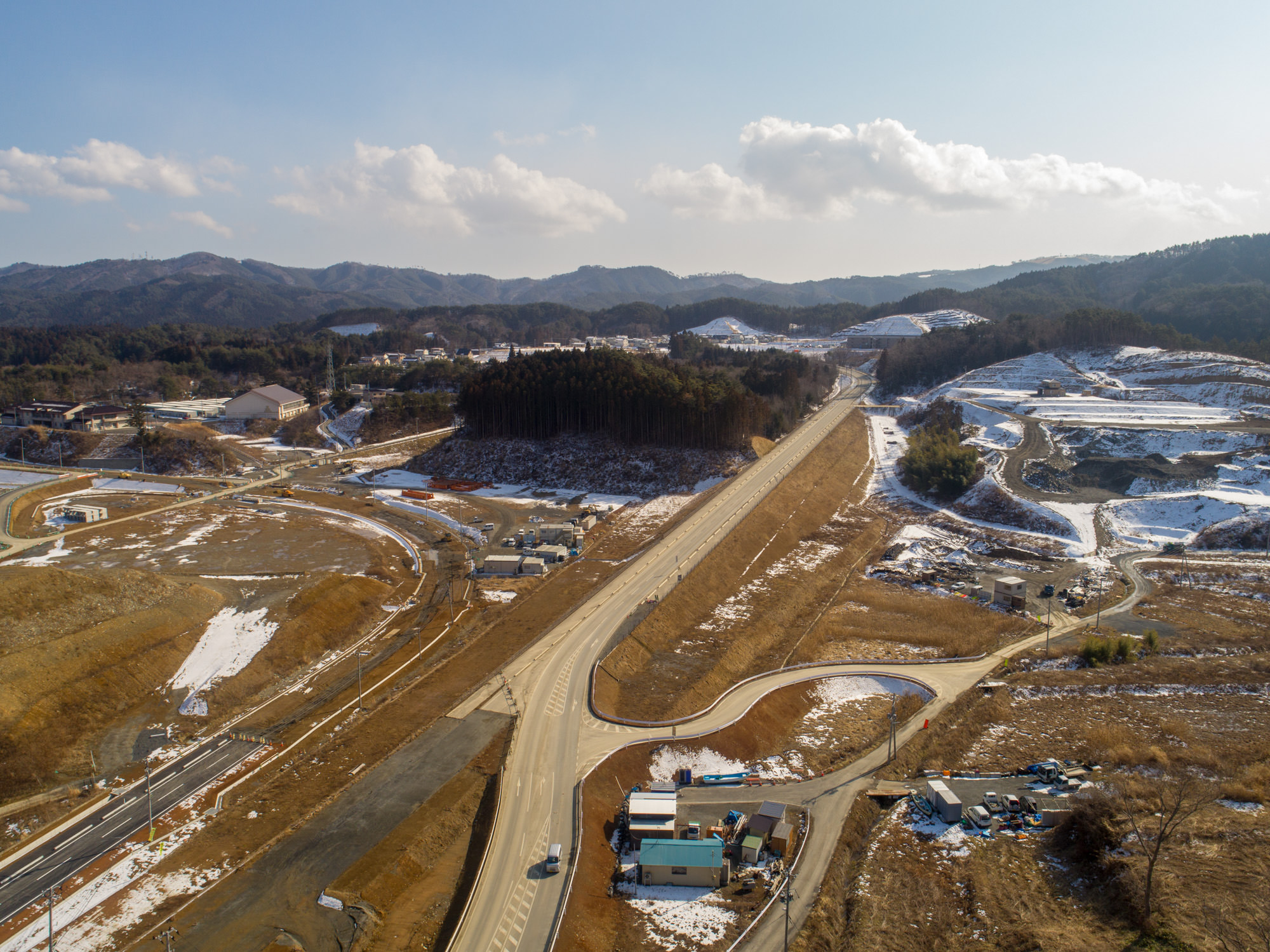 戸倉地区の風景を上空から撮影した写真で、中央には新しく整備されたと思われる道路が通り、周囲には雪が残る場所が点在して冬の季節を感じさせ、背景には丘や山が広がり、所々に建物や構造物があり、手前の建物付近には数台の車が停まっている風景の写真
