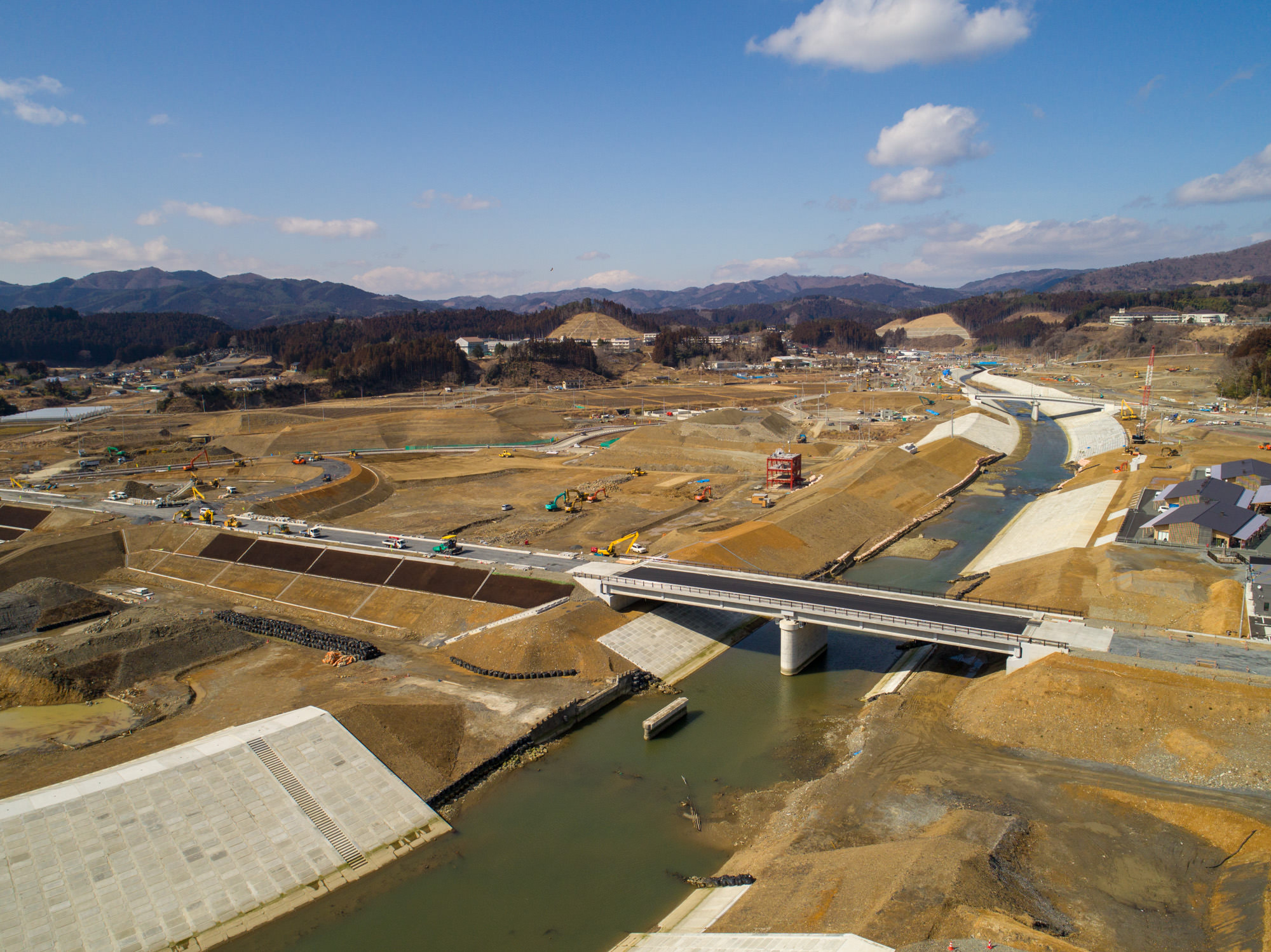 晴れ間のある空の下、中央を川が流れる大規模な建設現場を上空から撮影したもので、川には橋が架かり、現場には建設車両や機材が点在し、周囲には複数の道路や通路が整備されており、背景には丘や山々が広がる風景の写真