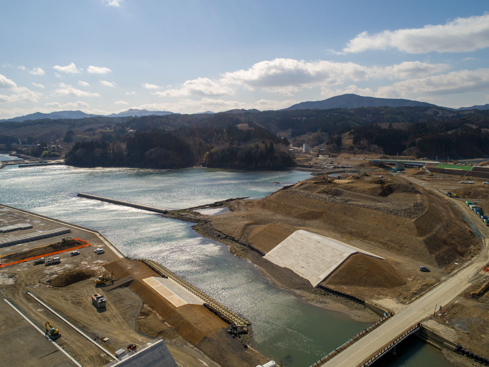 海沿いの地域で進行中の工事現場を撮影した写真で、岸辺には洪水対策や埋め立て工事と思われる大規模な土木作業やコンクリート構造物が建設されており、海へと流れる川には橋が架かり、背景には山や丘が広がり、現場には建設車両や機材が点在し、空は晴れて雲が少し浮かぶ工事現場の写真