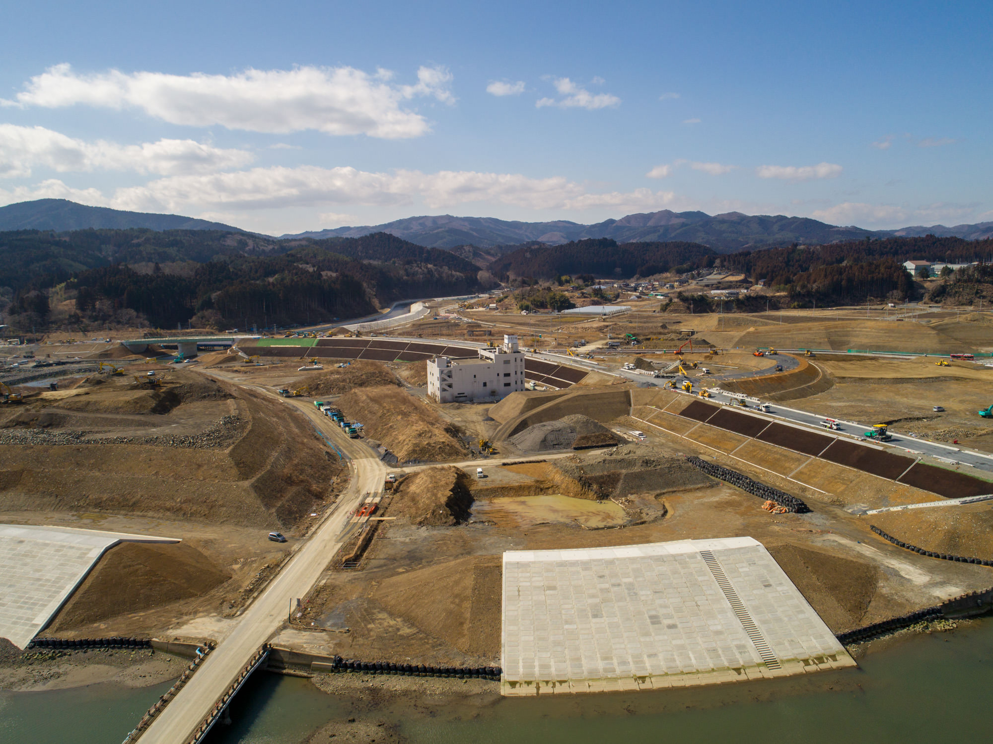 山々を背景にした地域の広大な工事現場を撮影した写真で、手前には川が流れ、両岸にコンクリート構造物が並び、橋がその上を横断し、中央には未完成の建物と思われる高台の構造物があり、現場内には建設車両や機材が点在し、草地や未舗装の道路が敷地内をつないでいる工事現場の写真
