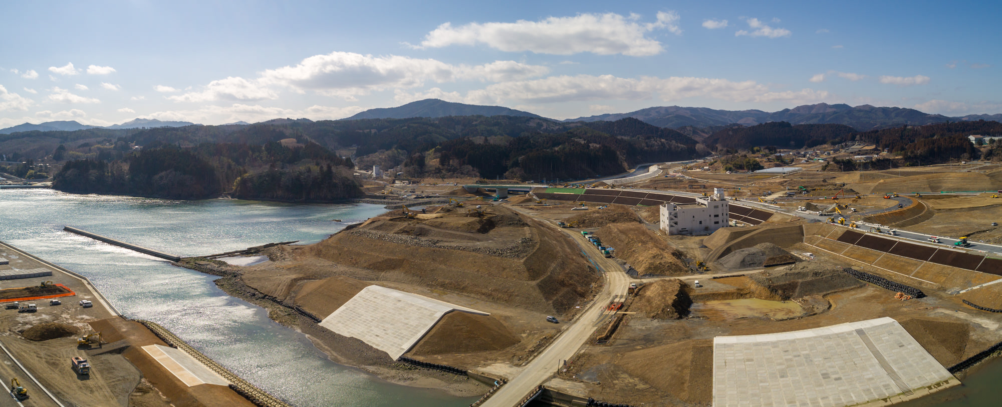 海へとつながる川の近くにある工事現場を広く見渡した写真で、周囲には山や丘が広がり、現場内には大型のコンクリート構造物や堤防、道路や通路が整備され、建設機械や車両が点在し、空は晴れて雲が少し浮かぶ天気の良い日の工事現場のパノラマ写真