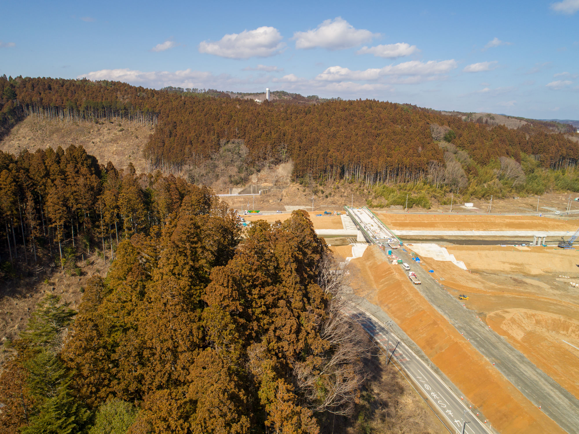晴れ間のある空の下、森林地帯にある道路を建設中の建設現場を上空から撮影したもので、複数の車両や機材が見られ、周囲は密集した木々に覆われ、背景には丘が広がる風景の写真