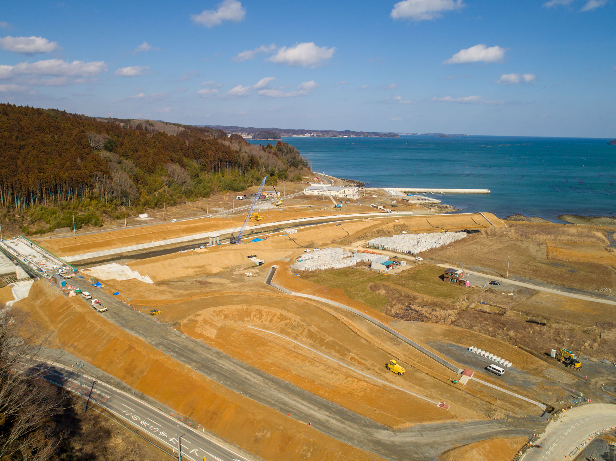 晴れ間のある空の下、海と森林に隣接する沿岸の建設現場を上空から撮影したもので、複数の道路や通路、構造物が整備中であり、現場にはさまざまな土木機械や車両が見られる風景の写真