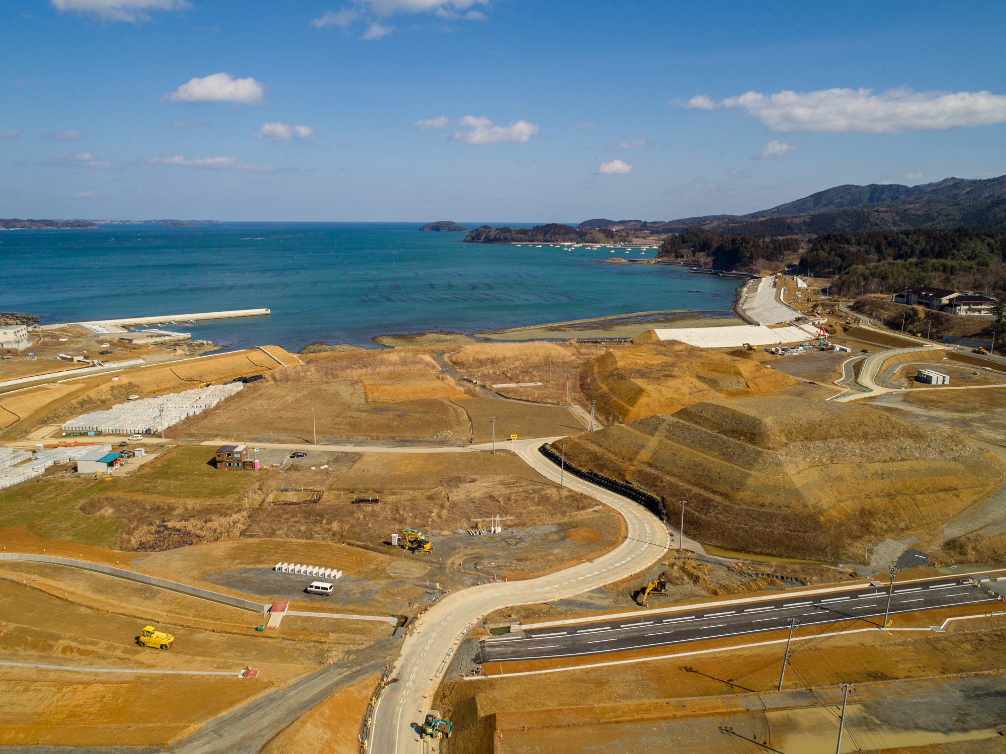 青空と散在する雲の下、右側に海と遠くの山々が広がる沿岸地域を上空から撮影したもので、複数の曲がりくねった新設道路やインフラが整備されており、建設車両が点在し、土地の一部が開発中と思われる風景の写真