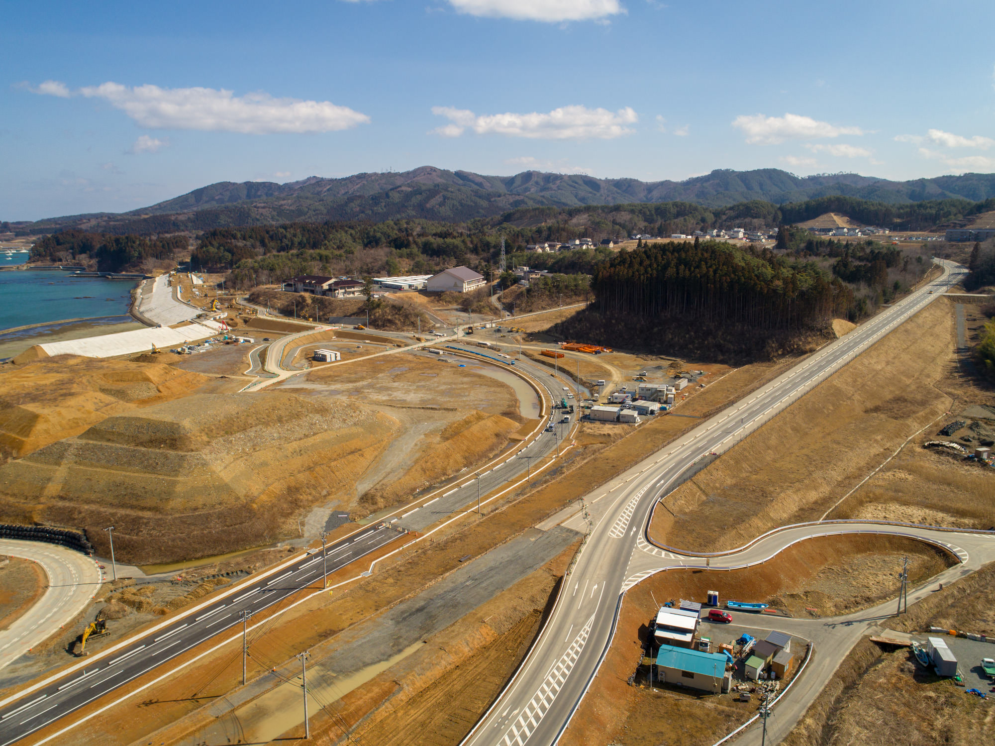 晴れ間のある空の下、左側に水辺が広がり、複数の道路が整備または建設中の地域を上空から撮影したもので、周囲には整地された土地や建物が点在し、背景には丘が広がる風景の写真