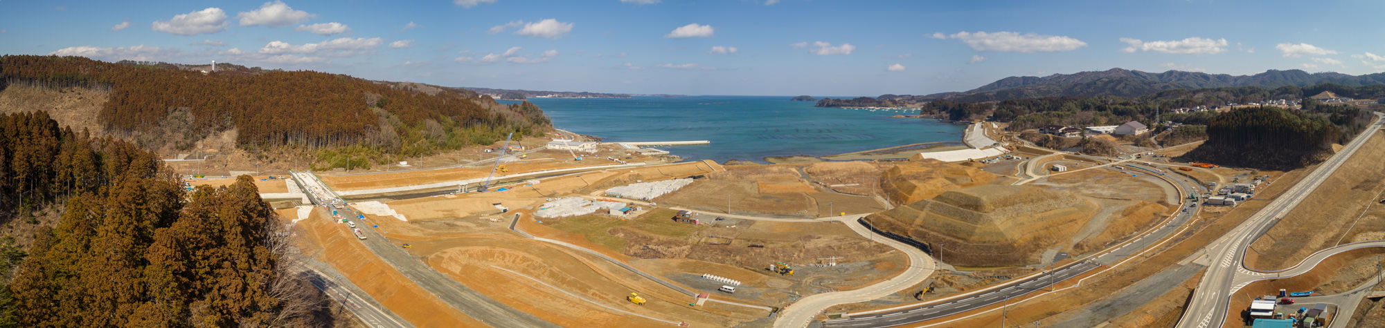 晴れ間のある空の下、海岸線と海を背景に、複数の曲がりくねった道路や建設車両、整地された土地が広がる開発または工事中の沿岸地域をパノラマで撮影したもので、左側には森林地帯、遠景には丘が見える風景の写真