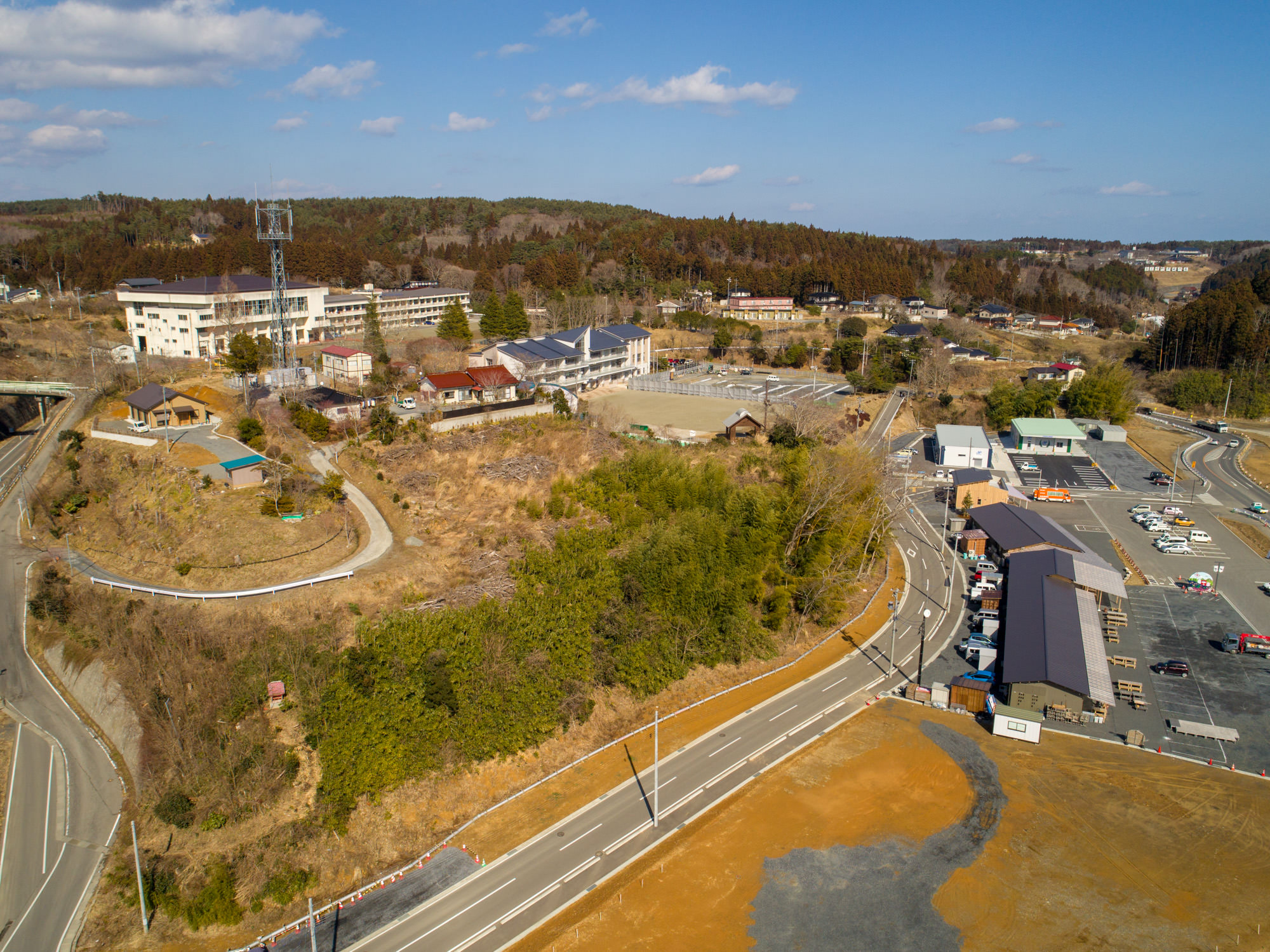 小さな町を上空から撮影した写真で、住宅や学校・事務所と思われる大きな建物、工場のような建物施設が点在し、道路が町をつなぎ、周囲には緑豊かな丘や森林が広がる風景の写真