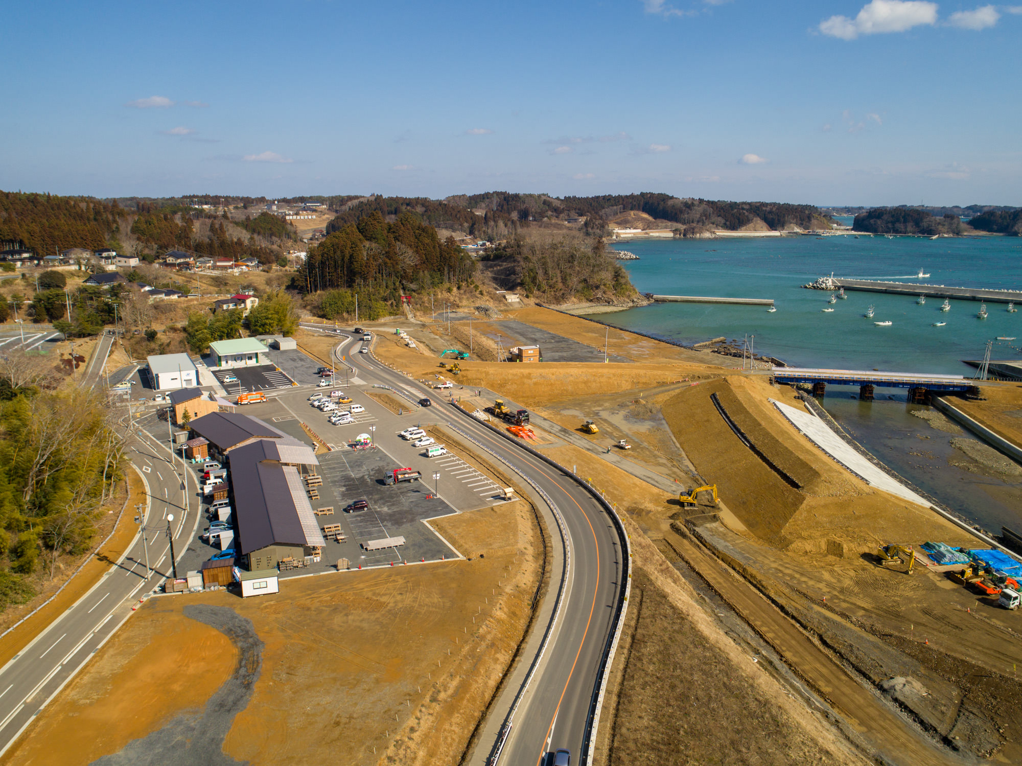 海沿いの地域を上空から撮影した写真で、中央に道路が通り、左側には建物や多くの車が停まっている駐車場があり、右側には桟橋に船が停泊する海が広がり、道路や水辺付近には工事作業や機材が見られ、背景には森林に覆われた丘と対岸の陸地が広がる風景の写真