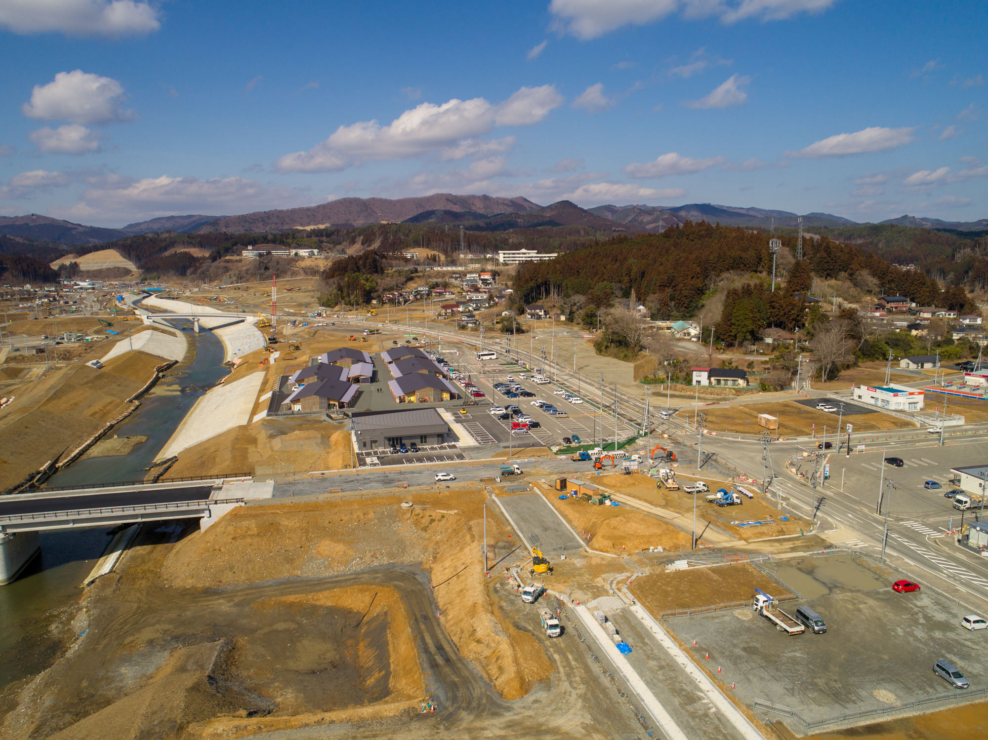 晴れ間のある空の下、左側に堤防で囲まれた川が流れる地域の建設現場を上空から撮影したもので、中央には車が見られる駐車場と商業施設の建物が複数あり、道路や交差点が広がり、一部は工事中で、背景には丘や森林が広がる風景の写真