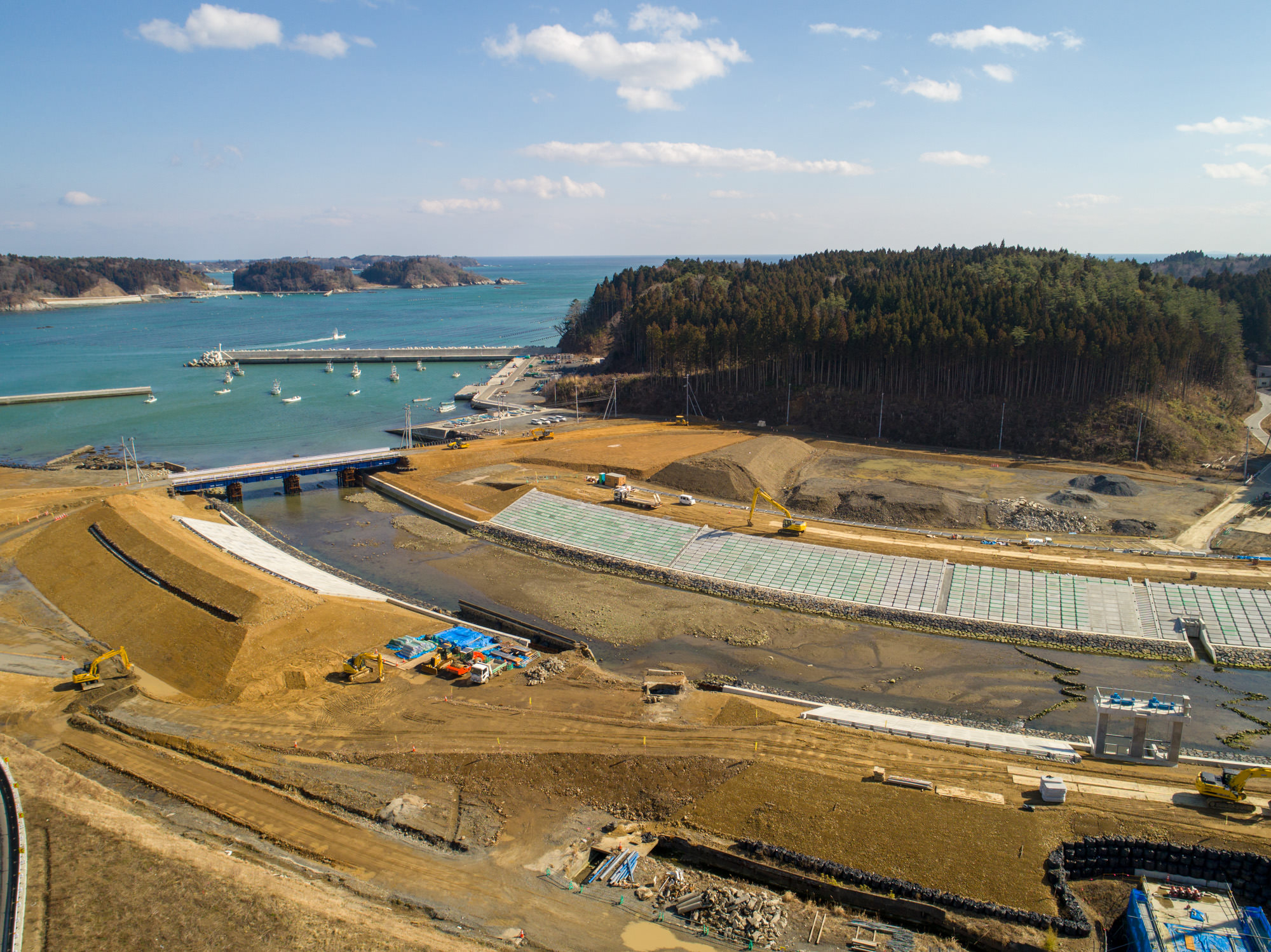 海へと流れる川沿いの工事現場を撮影した写真で、背景には船が停泊する桟橋付きの水辺があり、現場には段々に整地された広い土地が広がり、一部には緑色のマットや格子状の資材が敷かれ、複数のクレーンや重機が稼働しており、右側には森林に覆われた丘が見える工事現場の写真