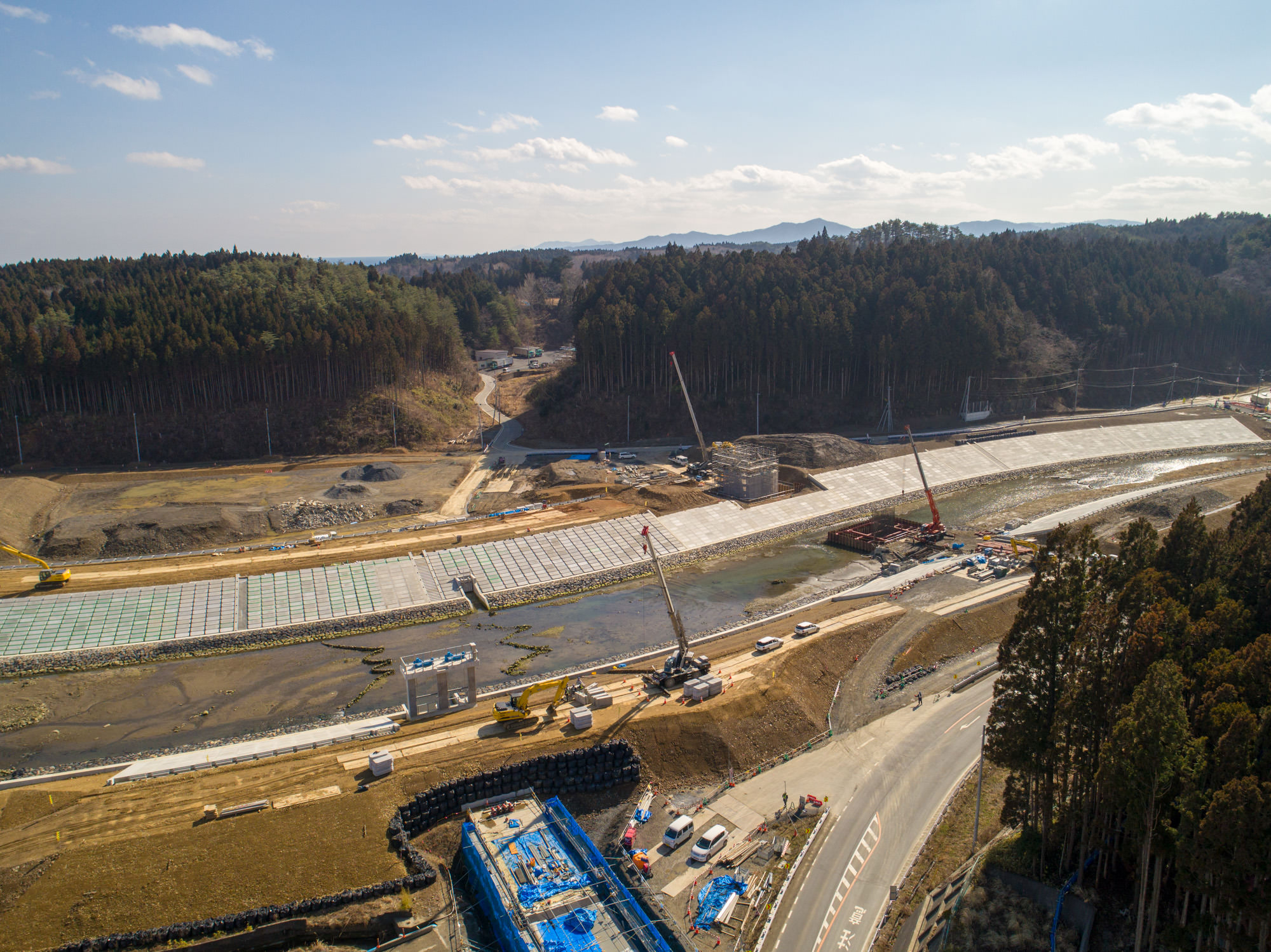 複数のクレーンや建設機材が稼働する広大な工事現場を撮影した写真で、現場には部分的にコンクリート板が敷かれた道路や橋の建設が進められており、周囲には森林に覆われた丘が広がり、地上には車両や作業員が見られ、空は晴れて雲が少し浮かぶ工事現場の写真