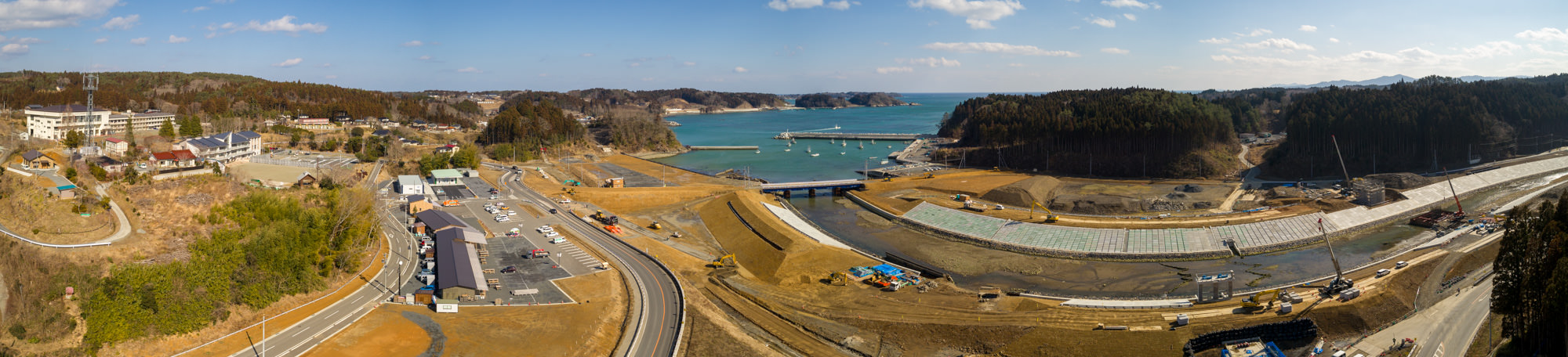 海沿いの地域を広く見渡したパノラマ写真で、左側には建物や複数の車が駐車している駐車場があり、中央には重機や機材が並ぶ工事現場があり、右側には川沿いで進行中の建設作業が見られ、クレーンなどの機材が配置され、背景には木々に覆われた丘と船が停泊する湾が広がり、空は晴れて雲が少し浮かぶ風景の写真