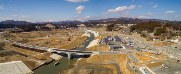 曇り空の下、中央を川が流れる建設現場を上空から撮影したもので、川には複数の橋が架かり、周囲には道路や建物が点在し、建設車両や機材が各所に見られ、背景には丘や山々が広がる風景の写真