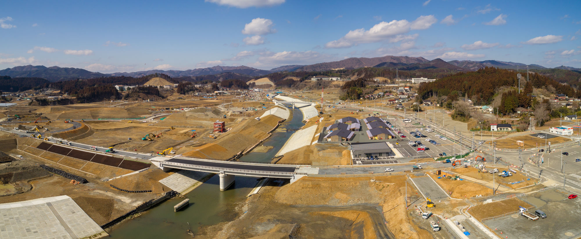 晴れ間のある空の下、中央を川が流れる建設現場をパノラマで撮影したもので、複数の橋が川を横断し、現場には建設車両や機材が点在しており、周囲には建物や道路、背景には丘が広がる風景の写真