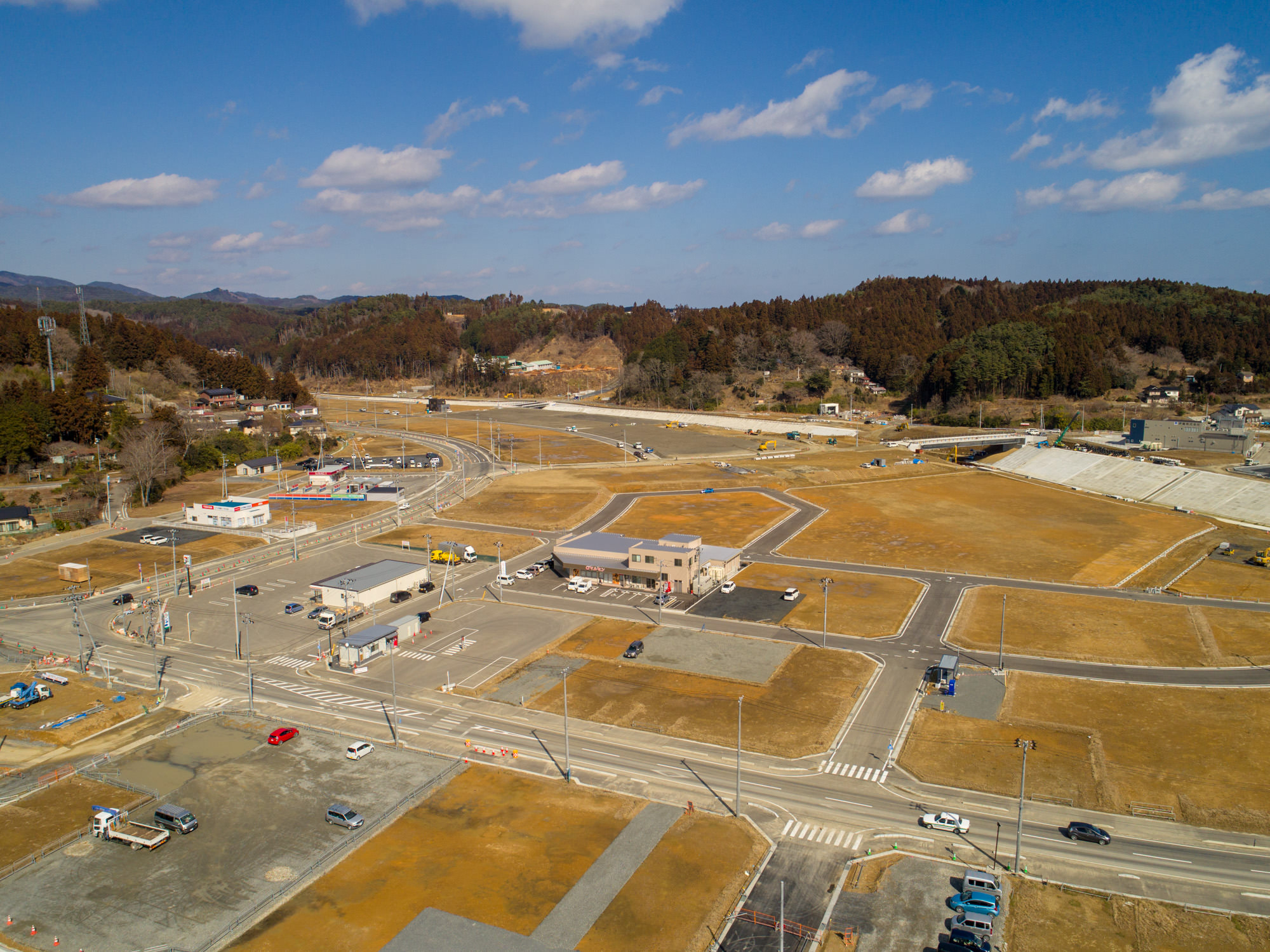 晴れ間のある空の下、複数の道路や建物、空き地が広がる地域を上空から撮影したもので、草地や舗装されたエリアが点在し、道路上には数台の車両が見られ、背景には木々に覆われた丘と点在する建物がある風景の写真