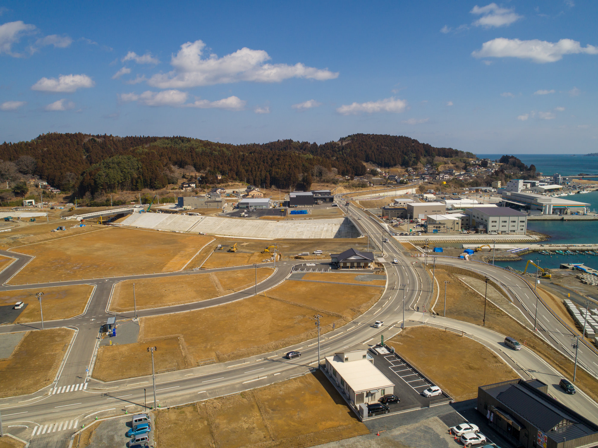 海沿いの地域を上空から撮影した写真で、複数の道路が交差し、建物や空き地が点在する開発途中の土地が広がり、背景には木々に覆われた丘と右側に海が見え、空は晴れて雲が少し浮かぶ風景の写真