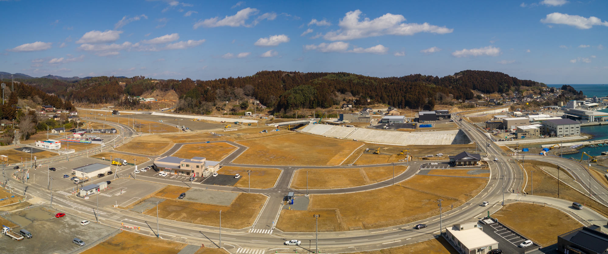晴れ間のある空の下、右側に海岸線が広がる沿岸の町を上空から撮影したもので、複数の道路が交差し、建物が点在する開発地と未開発地が混在しており、背景には森林に覆われた丘が見える風景のパノラマ写真