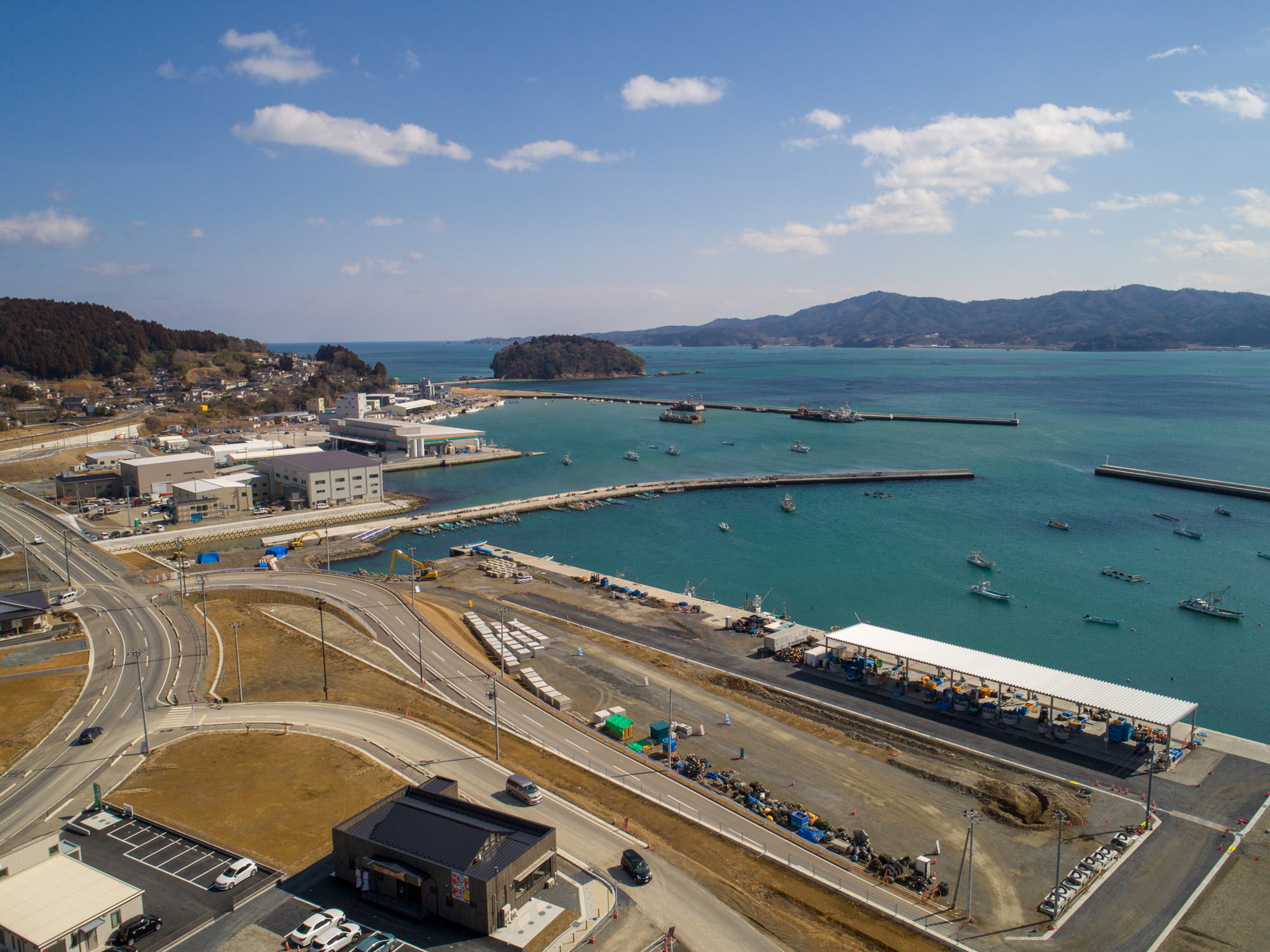 港のある海沿いの町を上空から撮影した写真で、湾内には複数の船が停泊し、桟橋や埠頭が海に突き出し、岸辺には道路や駐車場を含む建物や施設が並び、背景には山々が広がり、澄んだ青空が広がる風景の写真