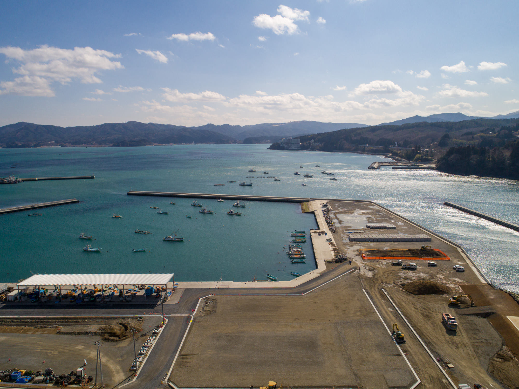 港のある海岸地域を上空から撮影した写真で、海には複数の船が停泊・航行しており、防波堤が波を防ぐように海に突き出し、周囲には工事現場や道路、建物があり、背景には山や丘が広がる風景の写真