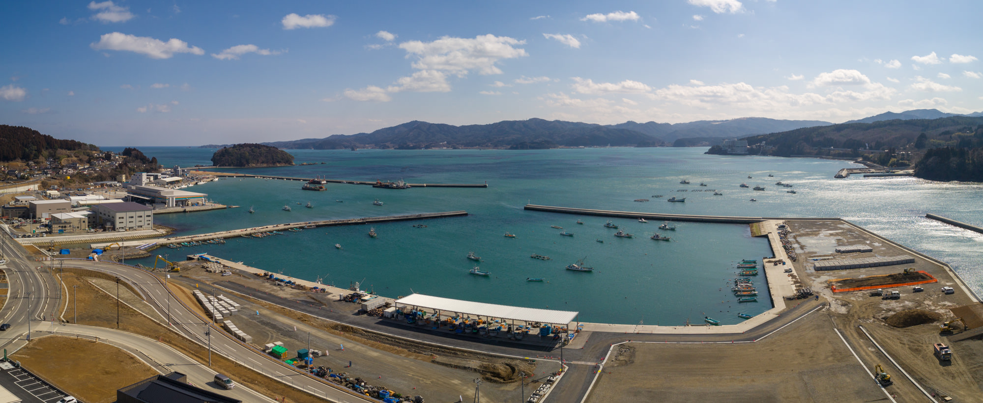 港のある海岸地域を広く見渡した写真で、海には複数の船が浮かび、防波堤が海に突き出し、岸辺には建物や施設が並び、背景には丘や山が広がる風景のパノラマ写真