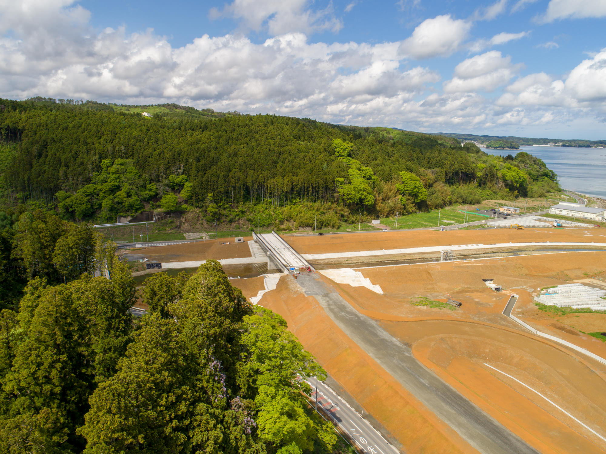 背景に密集した森林が広がり、中央には土地が整地され道路が敷かれた開発中の区域があり、右側には穏やかな水面の広がる海が見える、青空と雲が混ざった空の下で撮影された上空からの風景の写真
