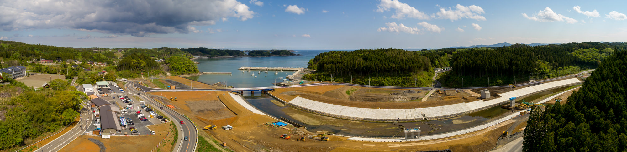 海や島々が背景に広がる海岸地域で、前景には土木作業や道路建設が進む工事現場が広がり、橋がその区域を横断して両側をつなぎ、複数の車両や重機、左側には建物や構造物が見られ、周囲には緑豊かな木々が広がる様子をパノラマで捉えた風景の写真