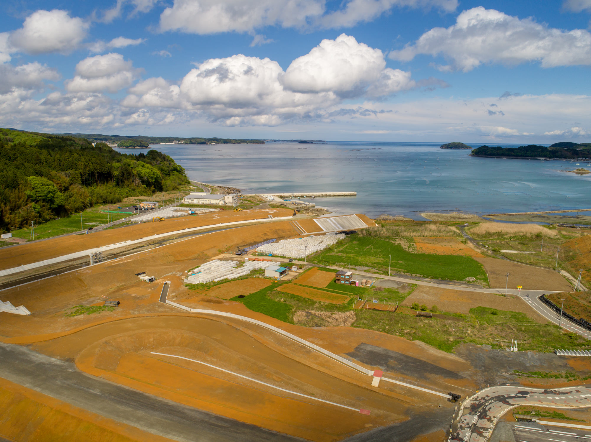 手前に緑の畑や開発中の土地が広がり、道路や建物が点在する海岸沿いの地域を写し、背景には広大な海と遠くの陸地が見え、空には青空と雲が混ざっている様子を上空から捉えた風景の写真