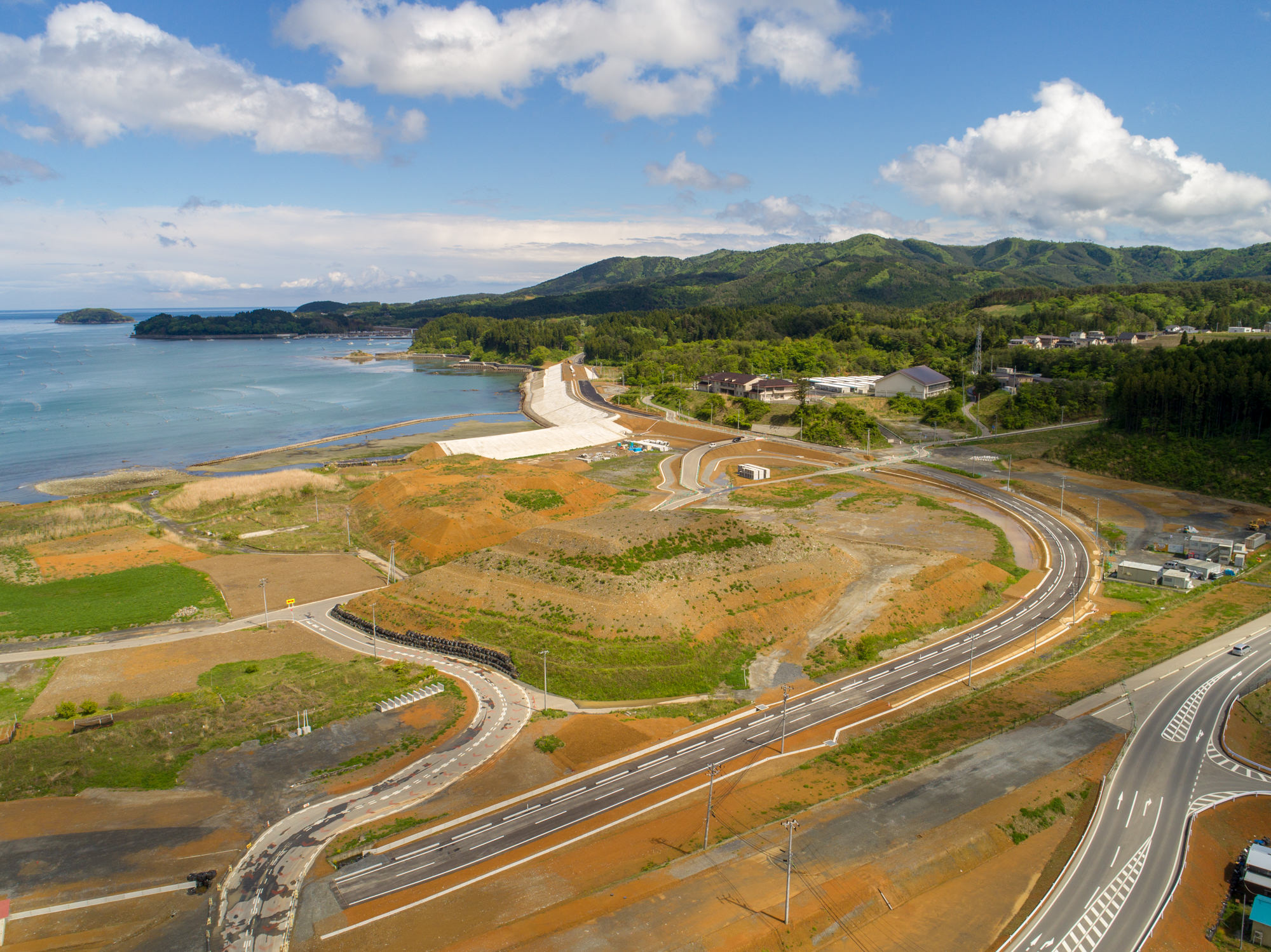 新しく建設された曲がりくねった道路が、開発中の土地を通って海岸へと続き、小さな浜辺が広がる沿岸部と、背景に緑豊かな丘や森林、その縁に建物が点在する様子を、晴れた空の下で上空から捉えた風景の写真