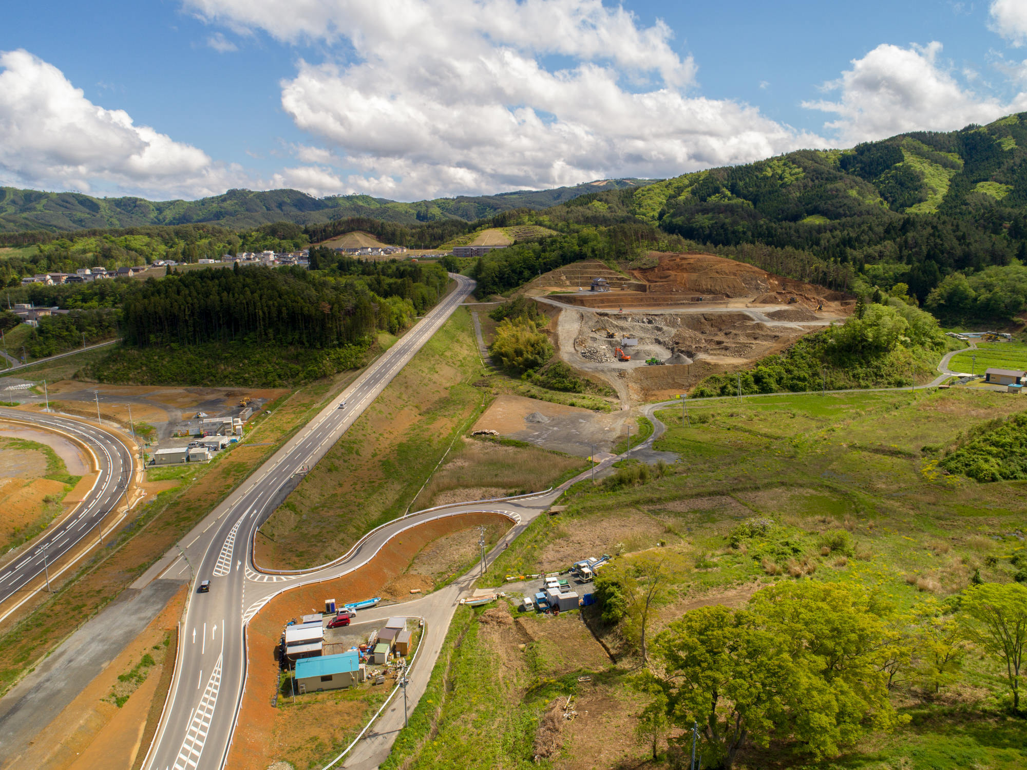 緑の丘が背景に広がる農村風景の中、道路が中央を走り、右側には工事や掘削作業が行われている区域があり、周囲には木々や草地、開けた畑、そして道路沿いに小さな建物や車両が点在する様子を上空から捉えた風景の写真
