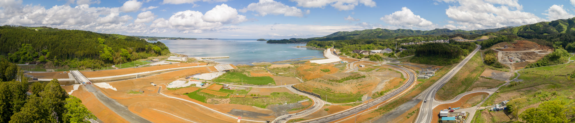 緑の多い丘陵地に沿って曲がりくねった道路が走り、所々に建設現場が点在する海沿いの地域をパノラマで捉え、穏やかな海と遠くに浮かぶ陸地、そして青空と雲が広がる空が一体となった風景の写真