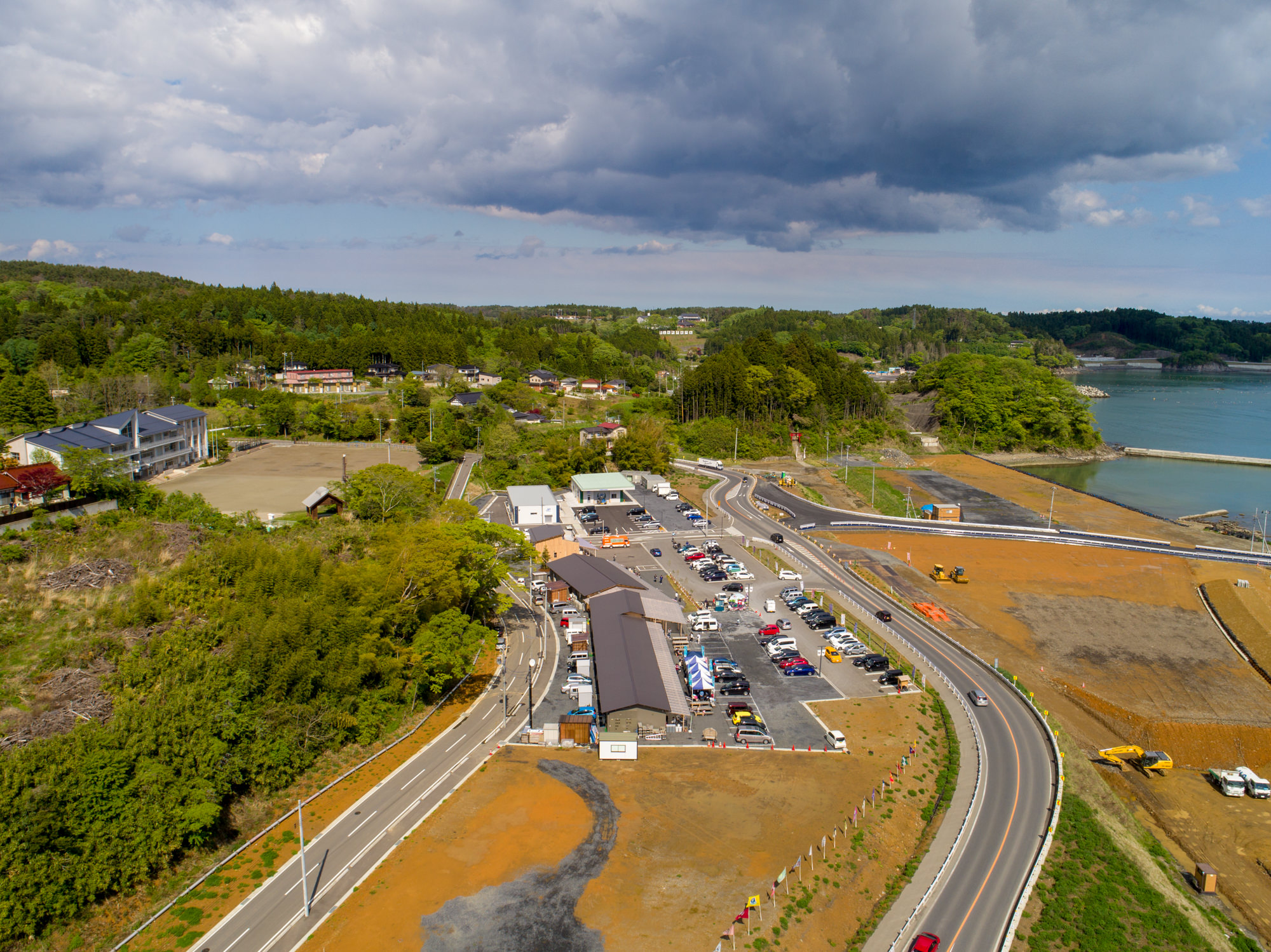 駐車場に多数の車が並び、周囲に建物や曲がりくねった道路が広がる小さな町を上空から捉えた写真で、右側には建設機材が見られ、背景には緑豊かな木々と水辺が広がり、空には雲が浮かぶ風景の写真
