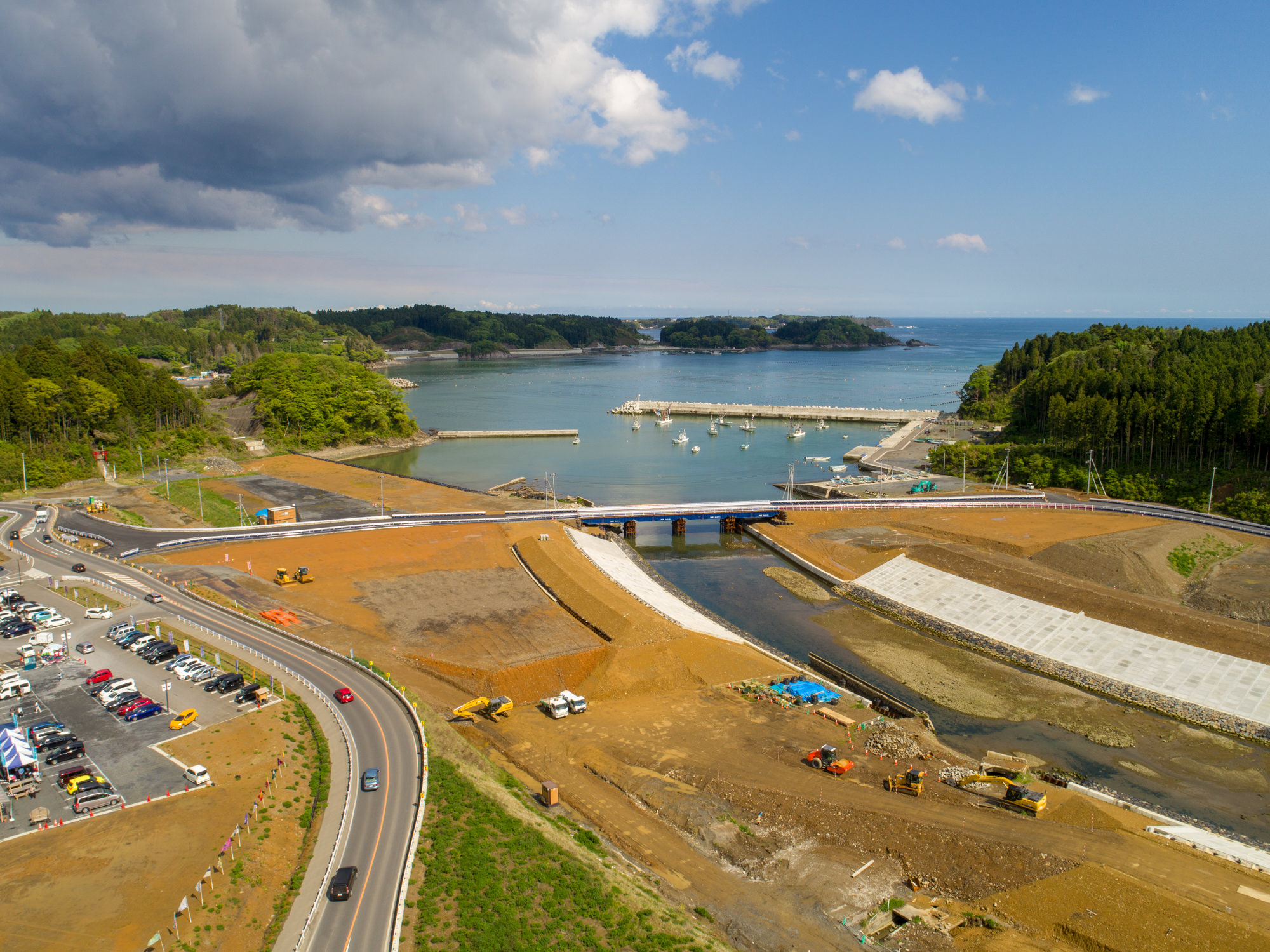 水路に架かる橋とその周辺で工事が進む海岸の建設現場を上空から捉えた写真で、現場にはショベルカーやトラックなどの建設車両が点在し、左側には駐車場と道路があり、背景には桟橋に停泊する船と広がる海が、雲が浮かぶ空の下に見える風景の写真