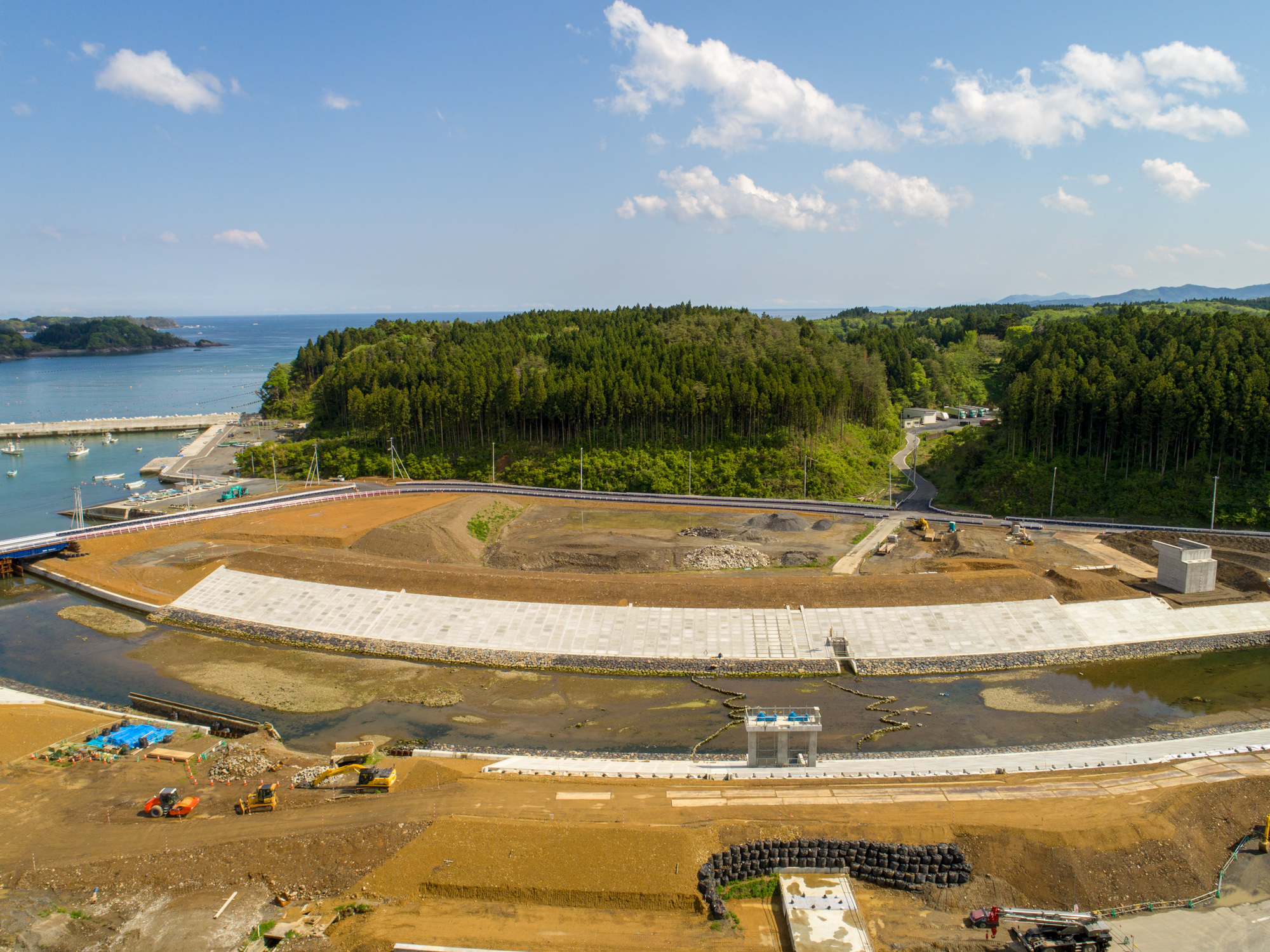 海に面した建設現場を上空から捉えた写真で、中央に流れる川沿いに大きく湾曲したコンクリート構造物があり、周囲には建設機材や資材、車両が点在し、背景には森林に覆われた丘と、遠くに海と小さな島々が広がる、雲が浮かぶ空の下の風景の写真