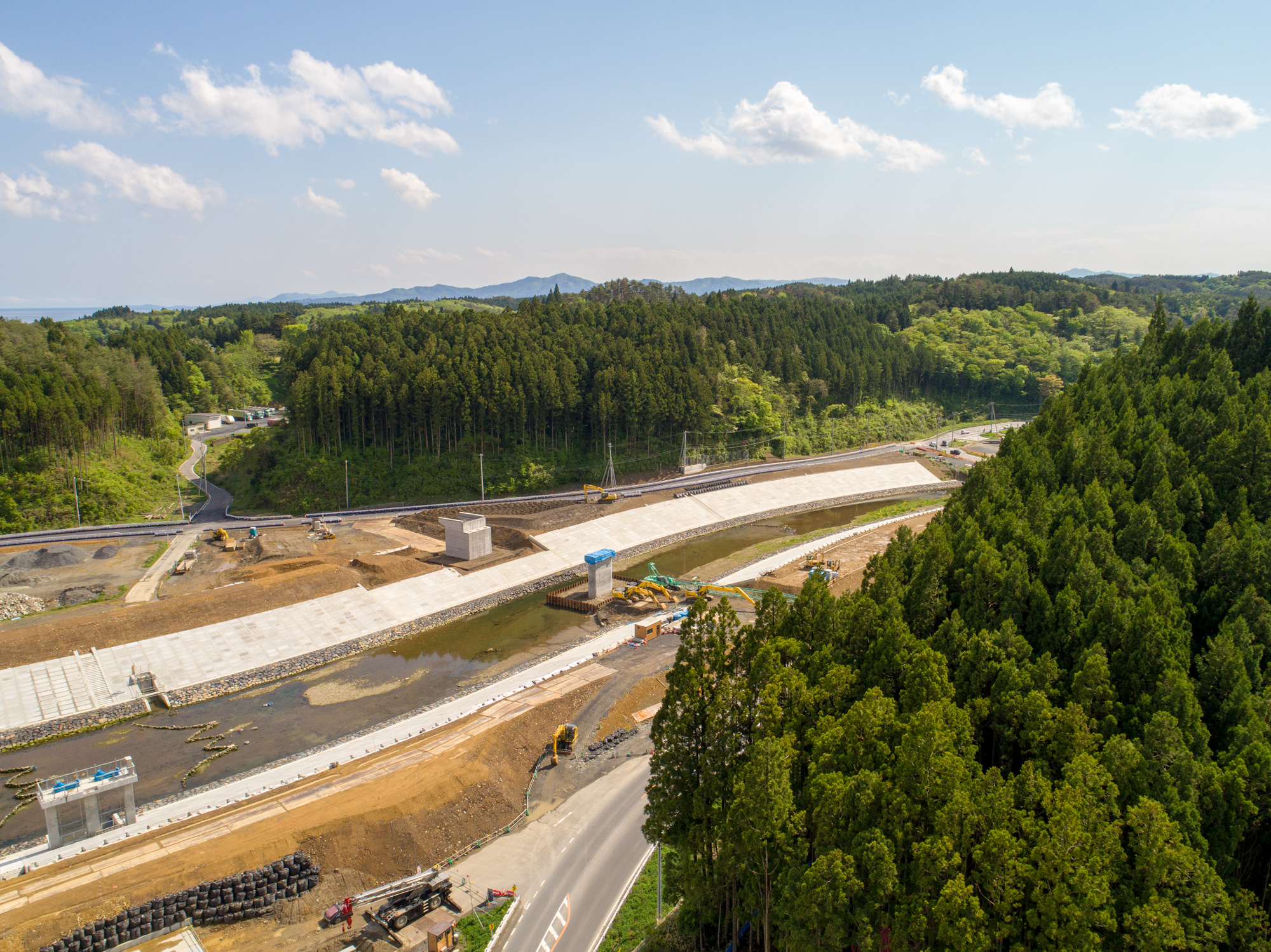森林と丘に囲まれた自然豊かな地域で、部分的に建設された橋が広がる工事現場を上空から捉えた写真で、現場には建設車両や機材、仮設の構造物が点在し、青空に雲が浮かぶ中でインフラ整備が進む様子が描かれた風景の写真