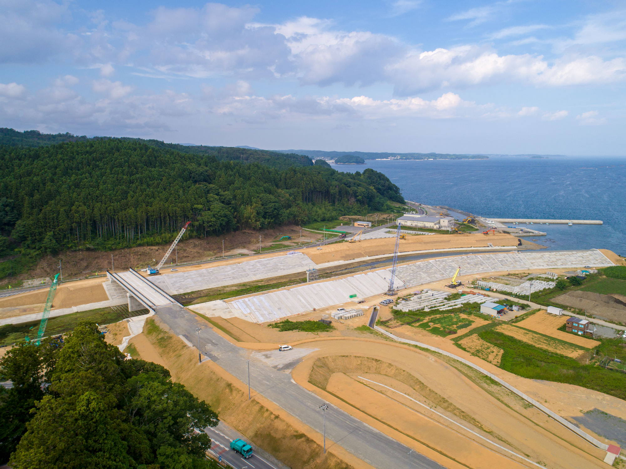 海に面した広大な工事現場で、複数のクレーンや車両が稼働し、森林に隣接した土地に道路や堤防、コンクリート舗装が整備されている様子を写した海岸開発の写真