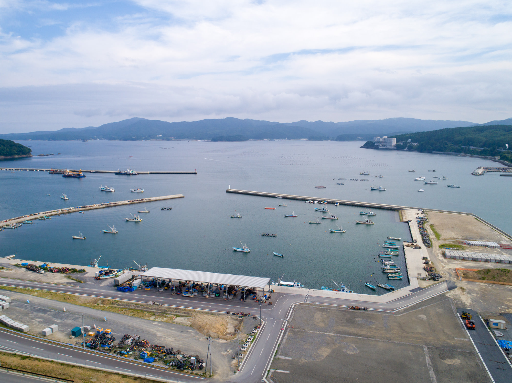 海に面した港町を上空から捉え、堤防に囲まれた港に多くの船が停泊し、沿岸には建物や道路が広がり、背景には緑に覆われた丘や山が見える様子を写した沿岸港湾の写真