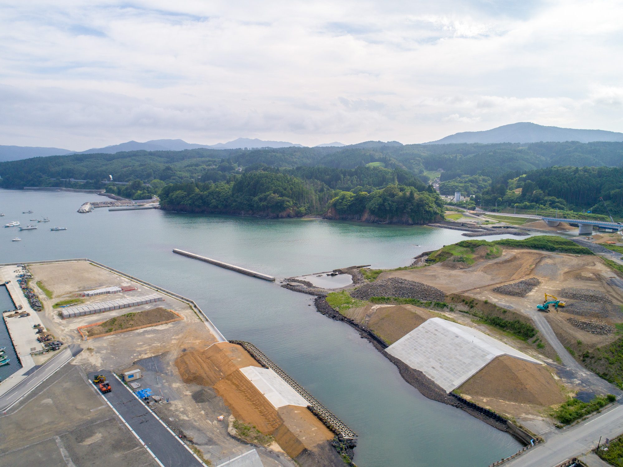 湾や入り江に面した沿岸地域を上空から捉え、道路や堤防、防波堤などのインフラ整備が進む工事現場と、背景に広がる森林に覆われた丘や山々が見える様子を写した海岸開発の様子の写真