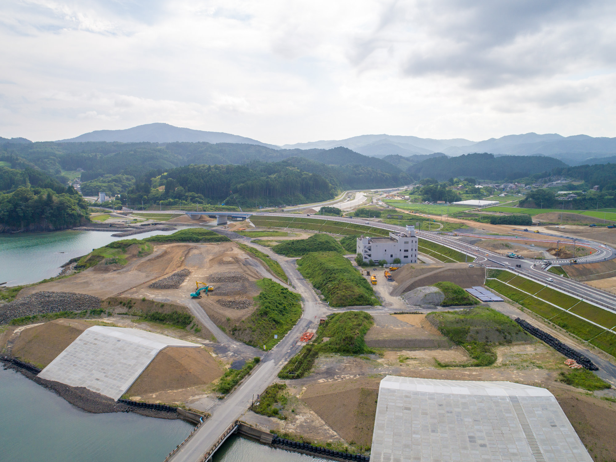 左側に水辺が広がる緑豊かな風景の中で、土の盛り土や建設車両、建設中の建物が点在し、道路や通路が整備されている様子を写した丘陵地の工事現場の写真