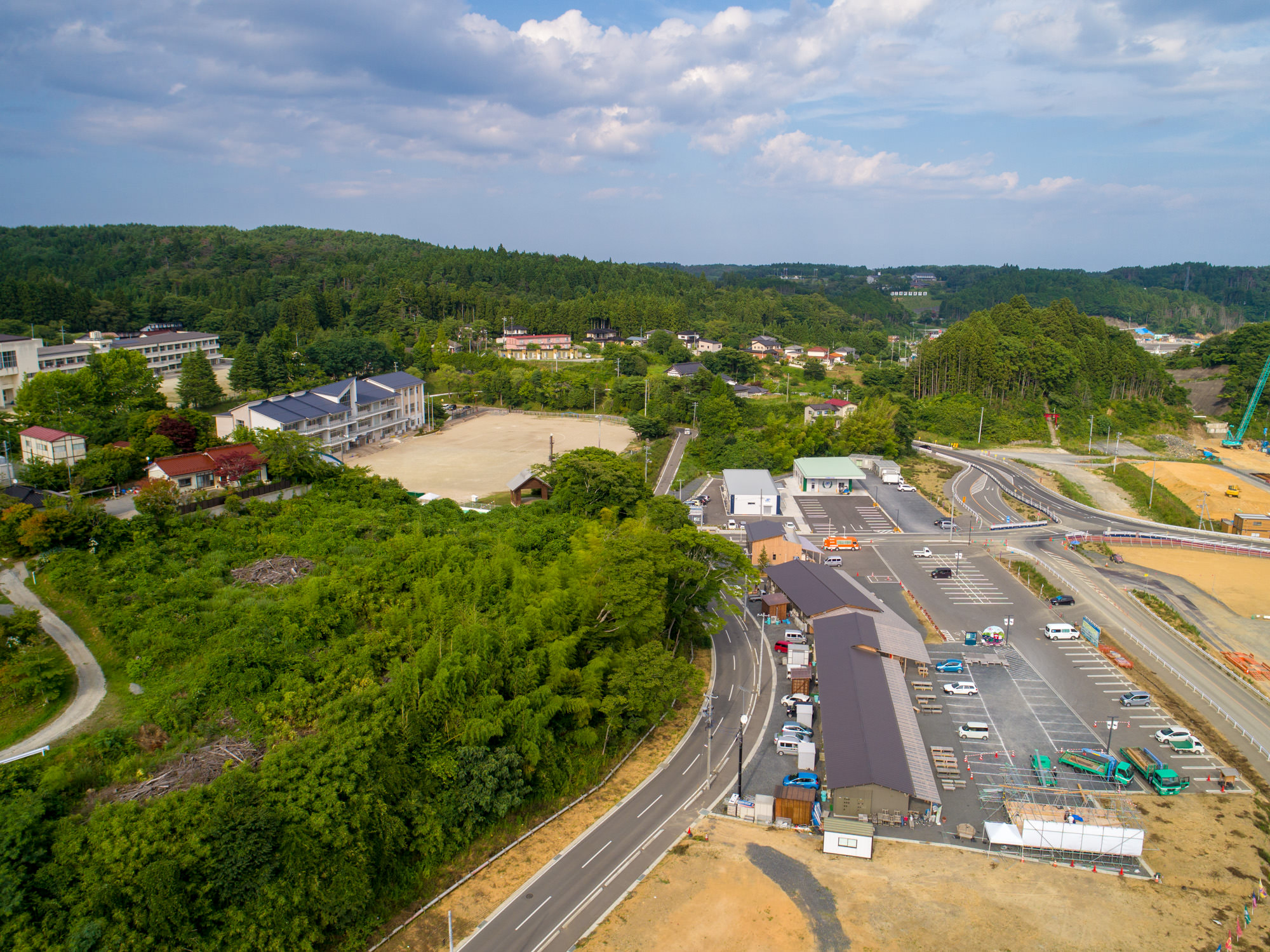 建物や道路、緑が混在する地域を上空から捉え、駐車場に複数の車が停まり、建設機械や住宅、学校または事務所と思われる建物が点在し、背景には森林に覆われた丘が広がる様子を写した歌津地区の写真