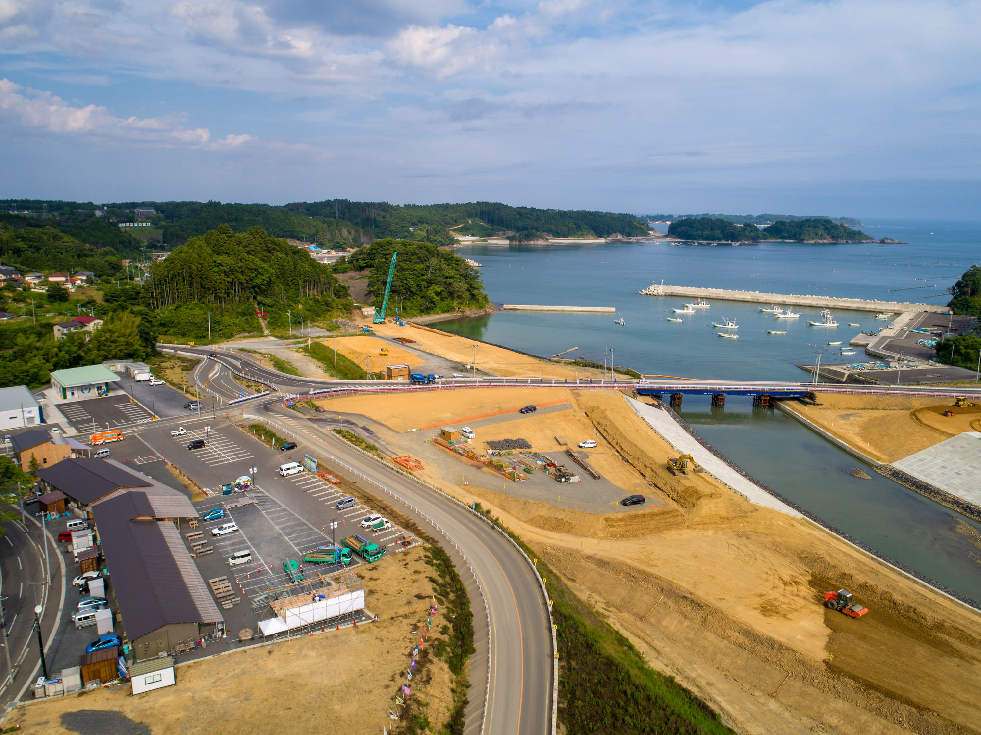 海に面した工事現場で、川にかかる橋や駐車場、複数の建物が広がり、クレーンや建設機械、停車中の車や港に停泊する船が見え、背景には木々に覆われた丘と水平線まで続く海が広がる様子を写した沿岸開発の写真