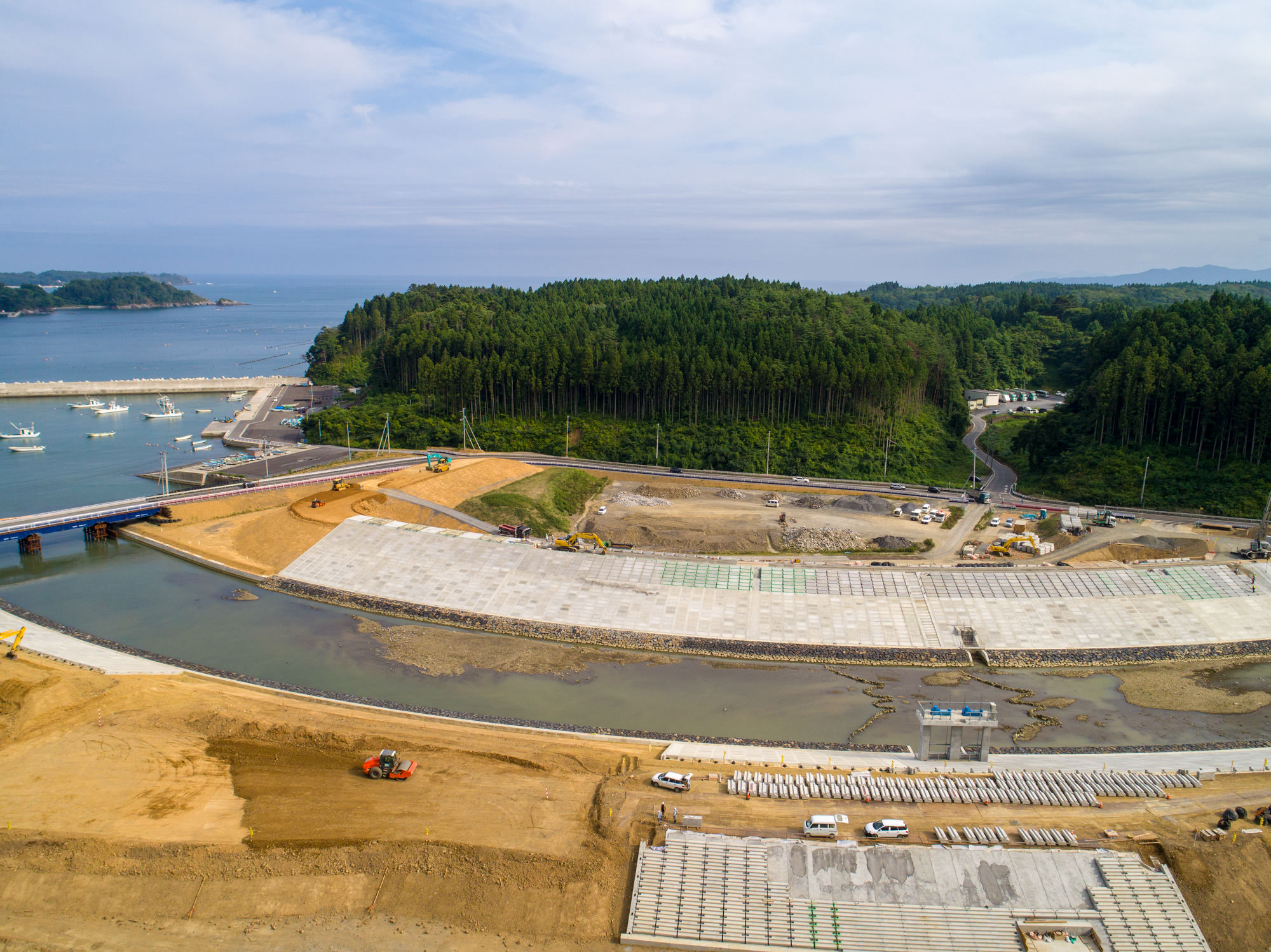 左側に海の見える地域で、中央に緩やかなカーブになった川が横断するように流れ、その両側にコンクリートの構造物が建設されている大規模な土木工事現場を上空から撮影した写真
