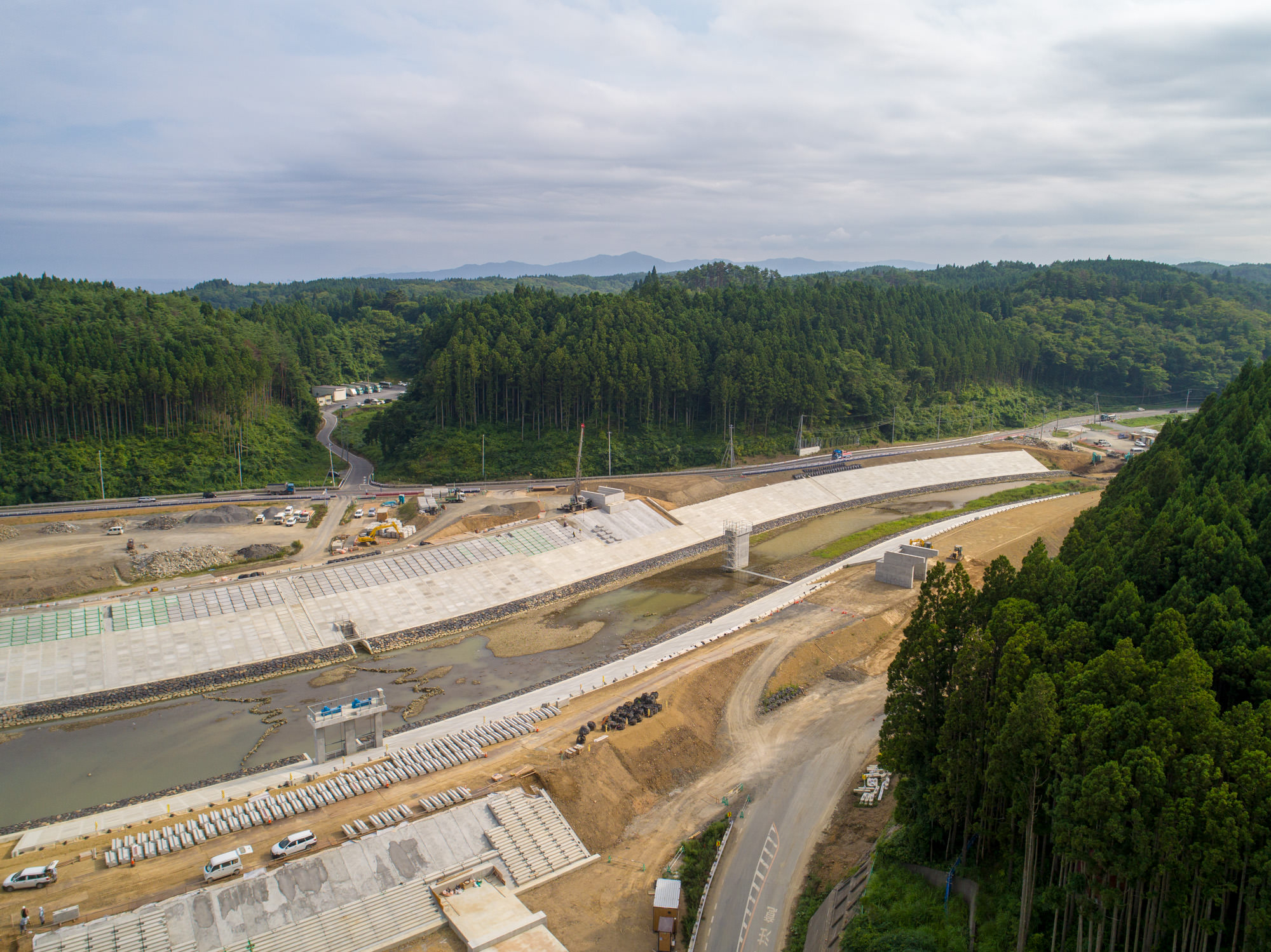 緑豊かな山々に囲まれた広い建設現場で、川沿いに大規模なコンクリート構造物が建設されている様子を上空から撮影した写真