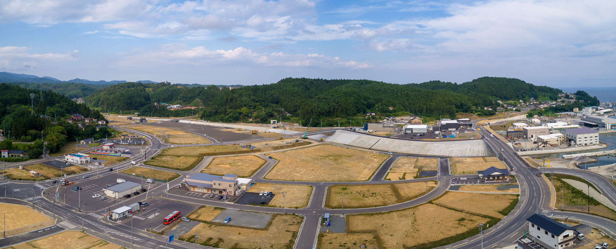 緑の丘を背景に、建物や道路、空き地が混在する半都市型の地域を上空から捉え、右側には工業・商業施設が見え、道路には車両が走る様子を写した志津川地区の風景のパノラマ写真