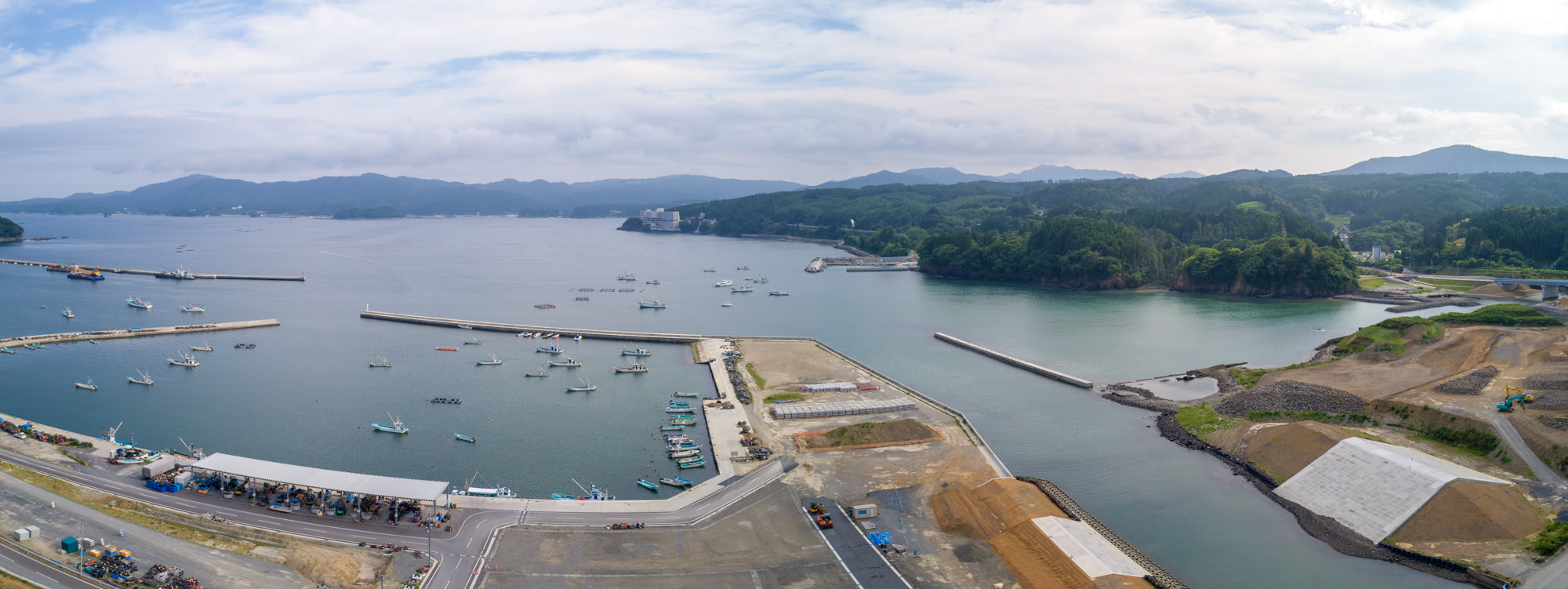 海に面した港町を上空から捉え、複数の船が停泊する港と海に突き出た防波堤、周囲の緑の丘や建物、工事中の道路や通路が広がる様子を写した沿岸地域のパノラマ写真