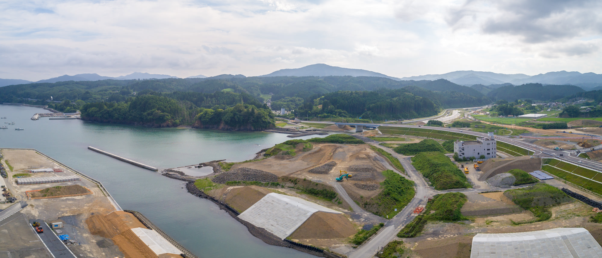 左側に海が広がる沿岸地域で、建設資材や機械が点在する工事現場が広がり、背景には丘や山々が見え、道路や通路が整備されている様子を写した海岸開発のパノラマ写真